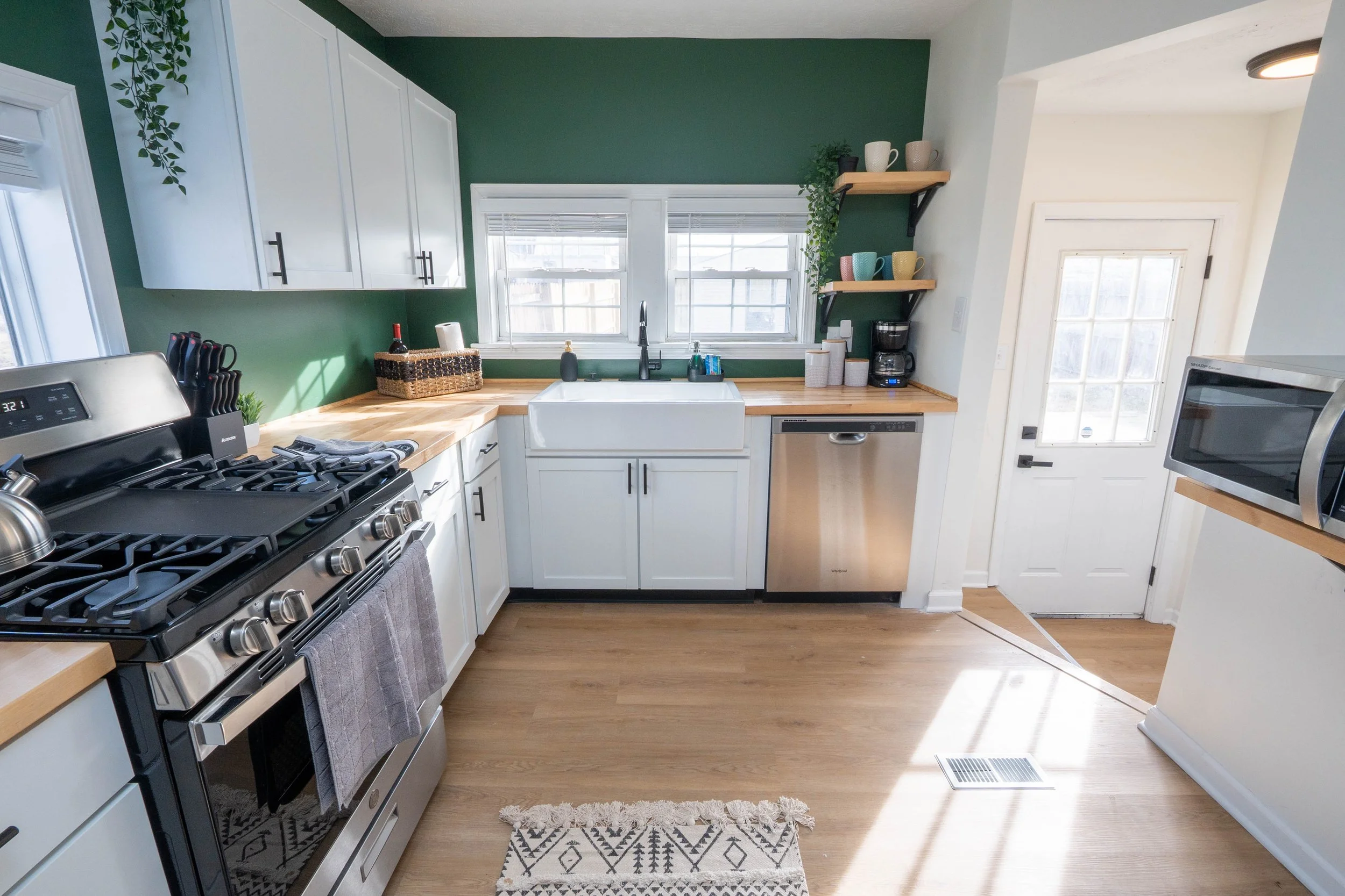 A modern kitchen with white cabinets, a green accent wall, wooden countertops, and various kitchen appliances including a stove, dishwasher, microwave, and coffee maker. There are windows above the sink, open shelves with cups, and decorative plants.