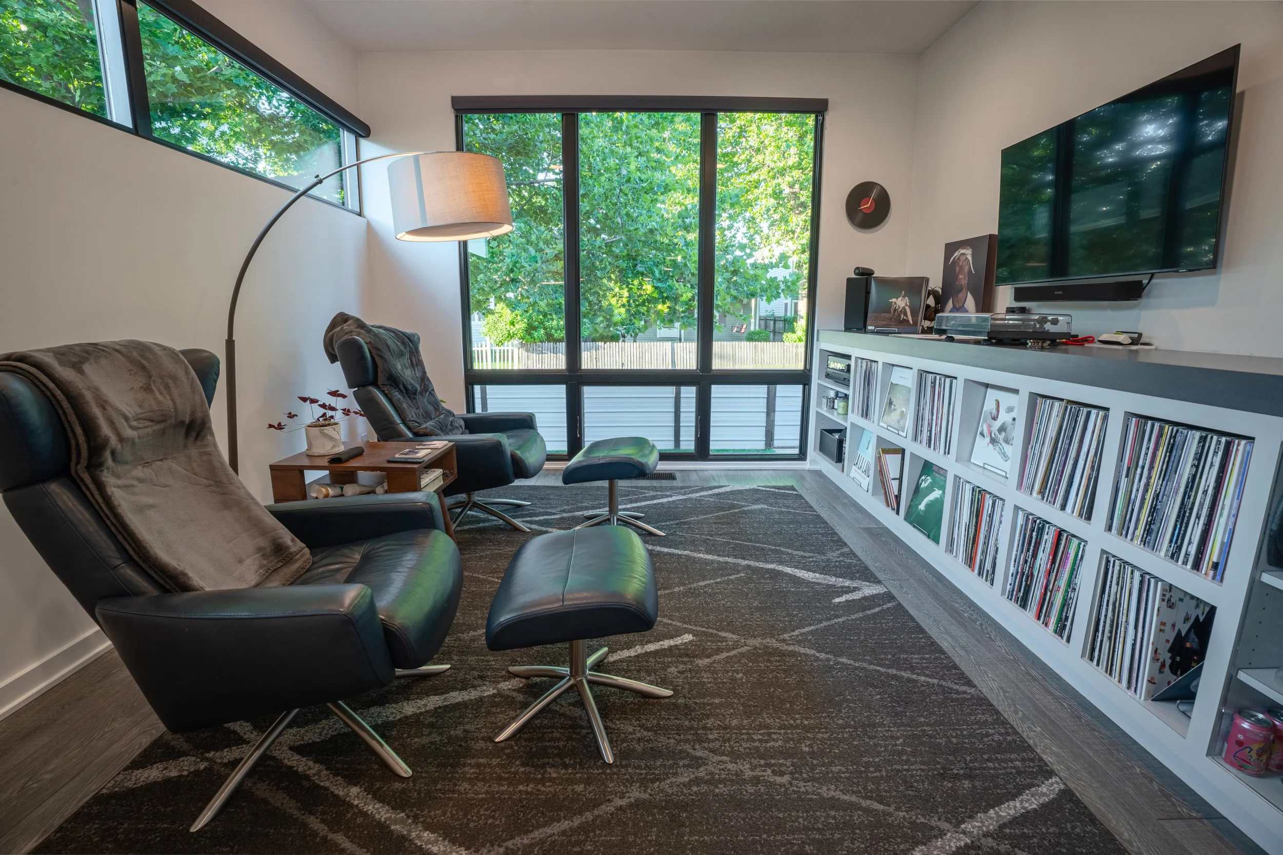 Living room with black leather chairs, a floor lamp, a white entertainment unit with books and records, a wall-mounted TV, large windows with green trees outside, and a patterned dark rug.