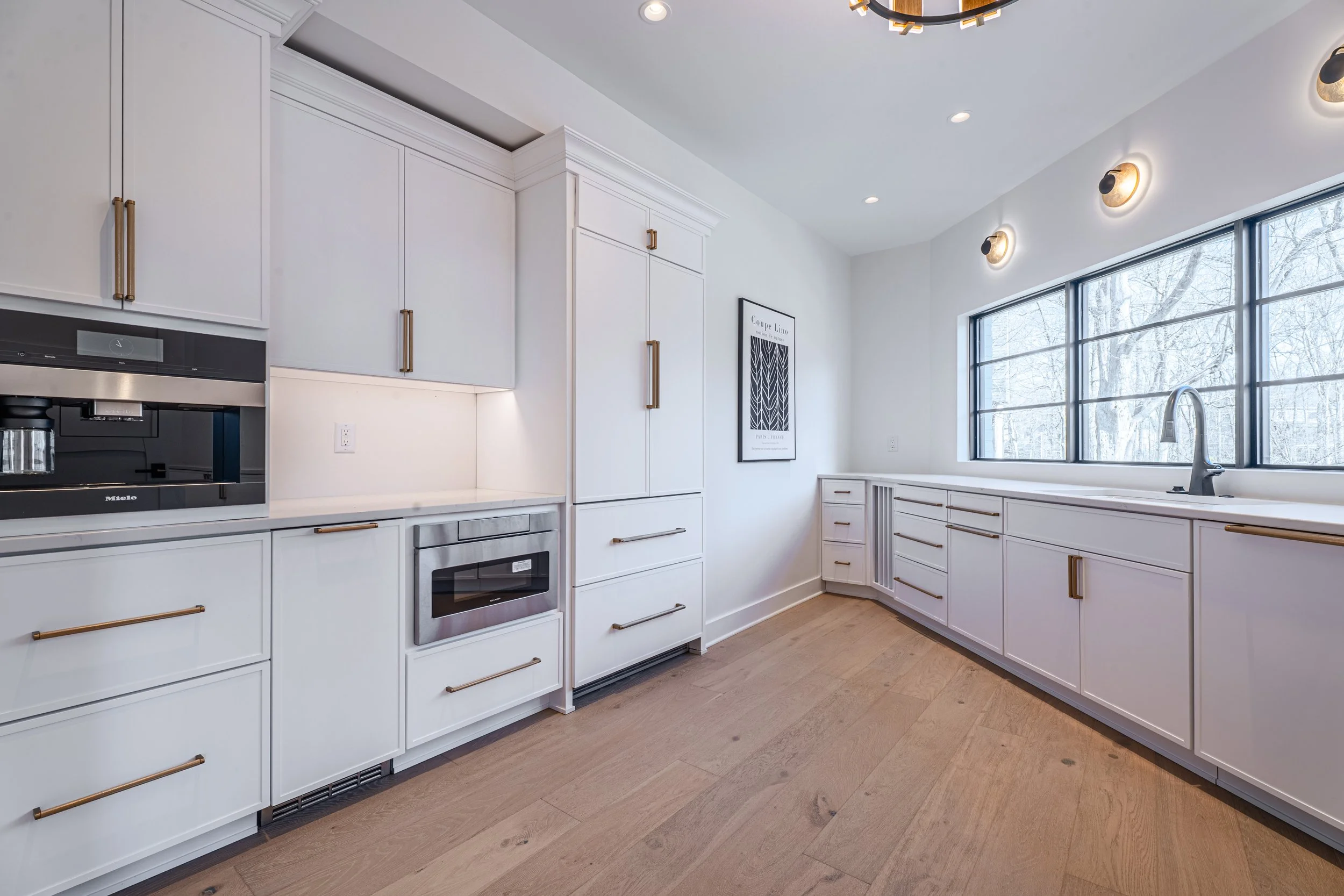 A modern kitchen with white cabinets, gold handles, stainless steel appliances, a large window, and light wood flooring.