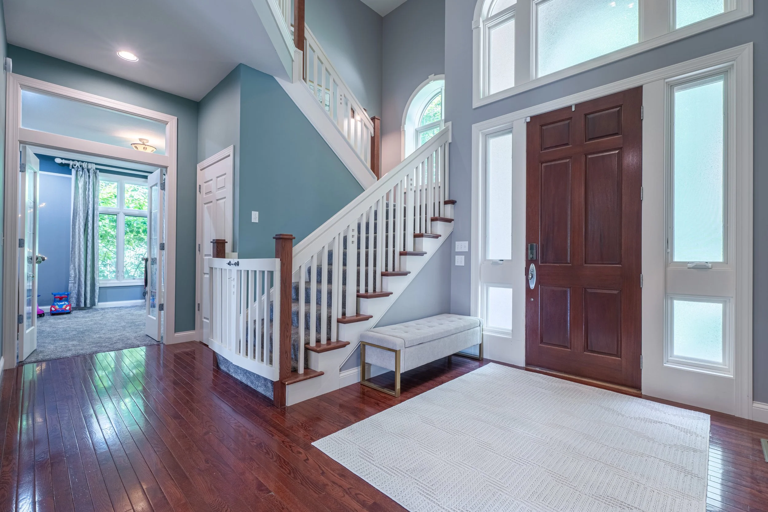 Interior of a home entryway with a staircase, a large wooden front door, and multiple windows letting in natural light. There's a small bench with a cushion near the door, and a hallway leading into another room with windows and children's toys.