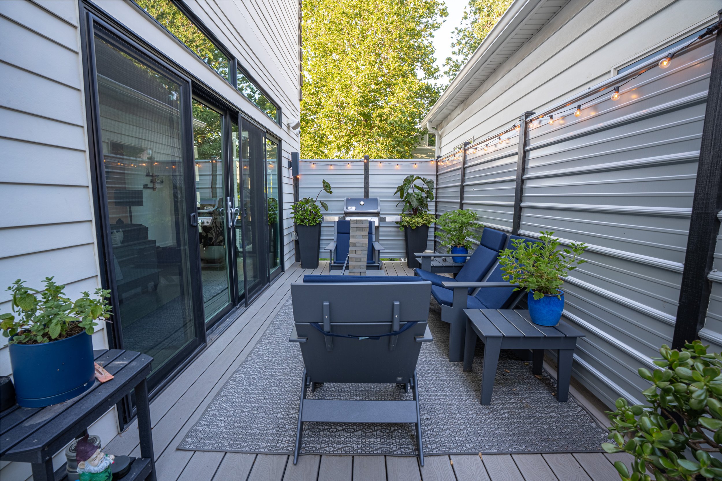 A small outdoor patio with gray furniture, potted plants, and string lights, enclosed by gray fencing and adjacent to a house with sliding glass doors.