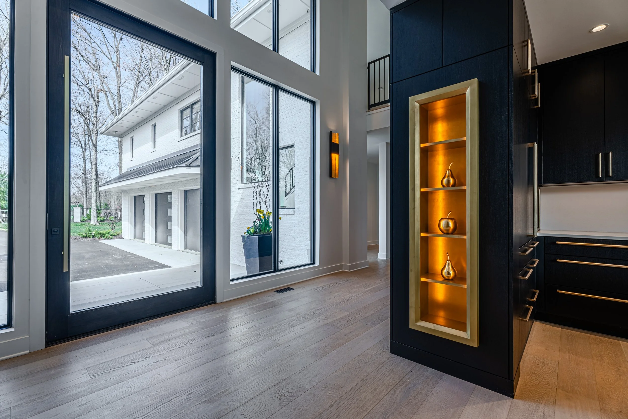 Interior view of a modern kitchen with large windows, a black cabinet with gold accents, and decorative gold vases on a shelf, with an outdoor view of a house with white siding and leafless trees.