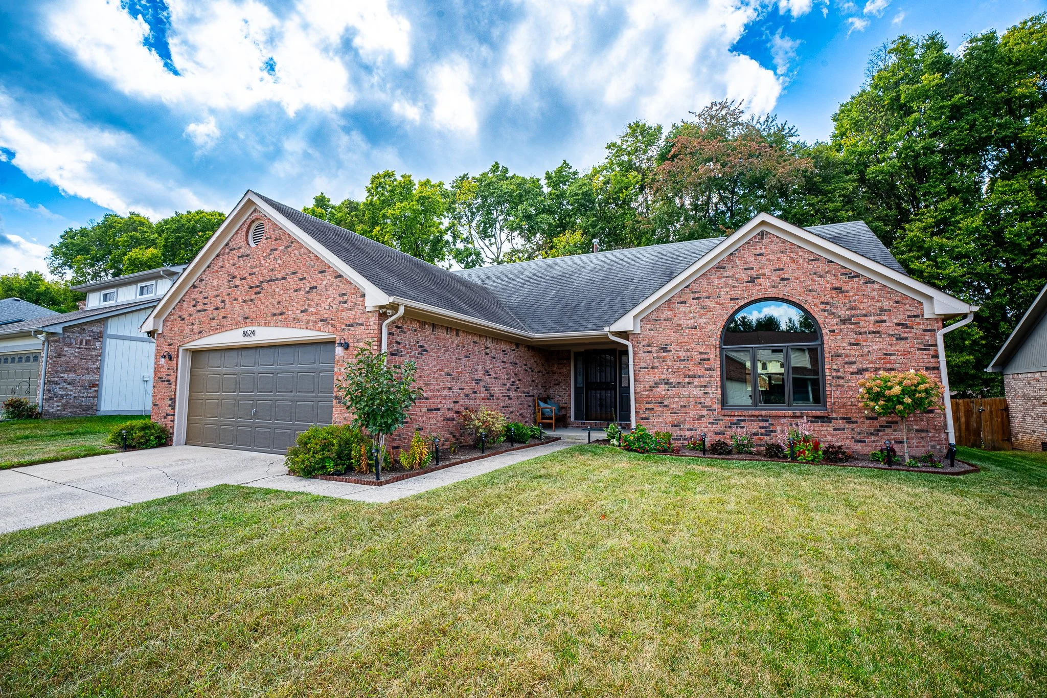 A brick house with a gray garage door, arched window, front porch, and lush green lawn under a partly cloudy sky.
