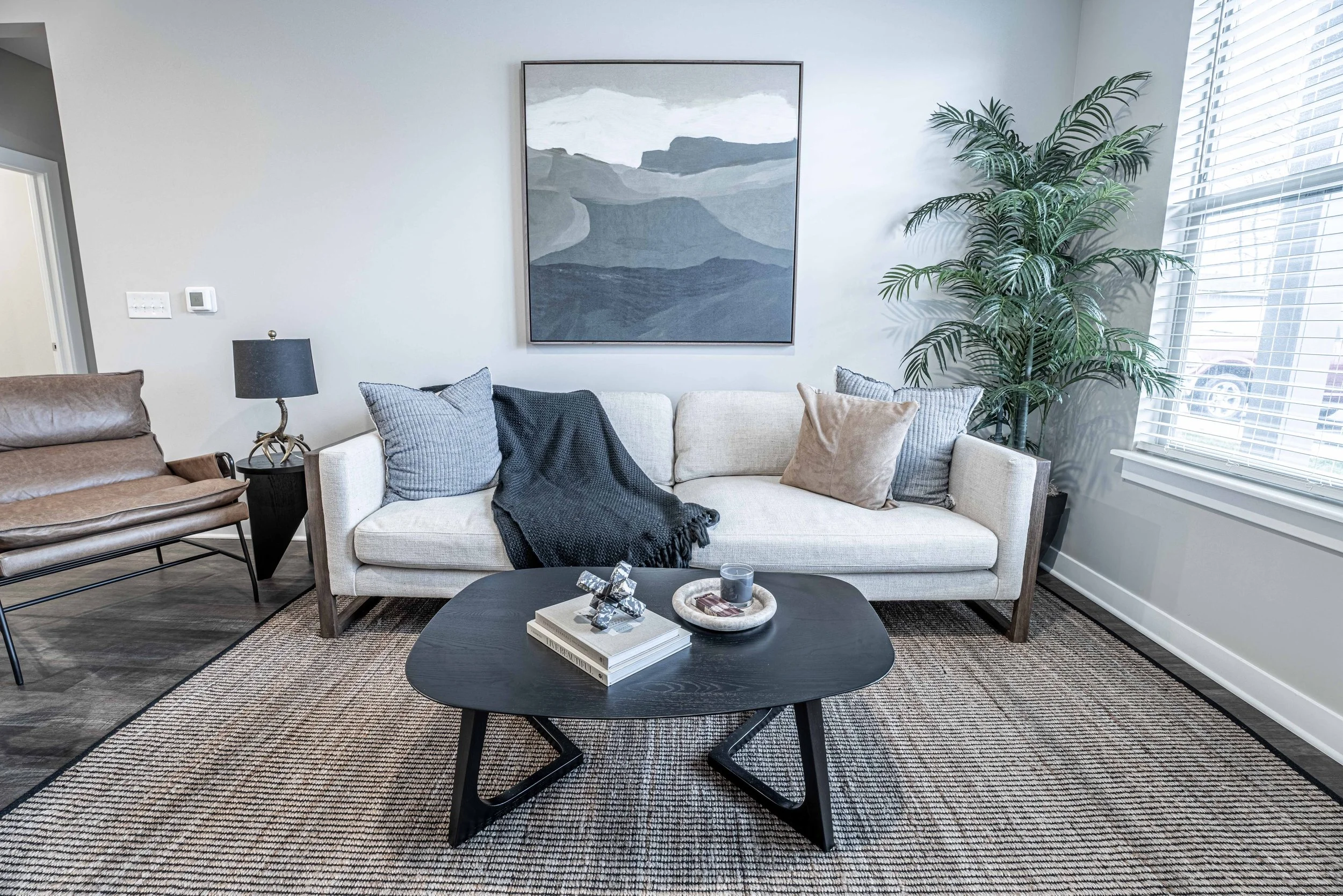 Living room with a beige sofa, dark wooden coffee table, blue pillows, and a black throw blanket. There is a large abstract mountain artwork on the wall, a tall potted plant near a window with white blinds, a black side table with a grey lamp, and a brown leather armchair on the left.