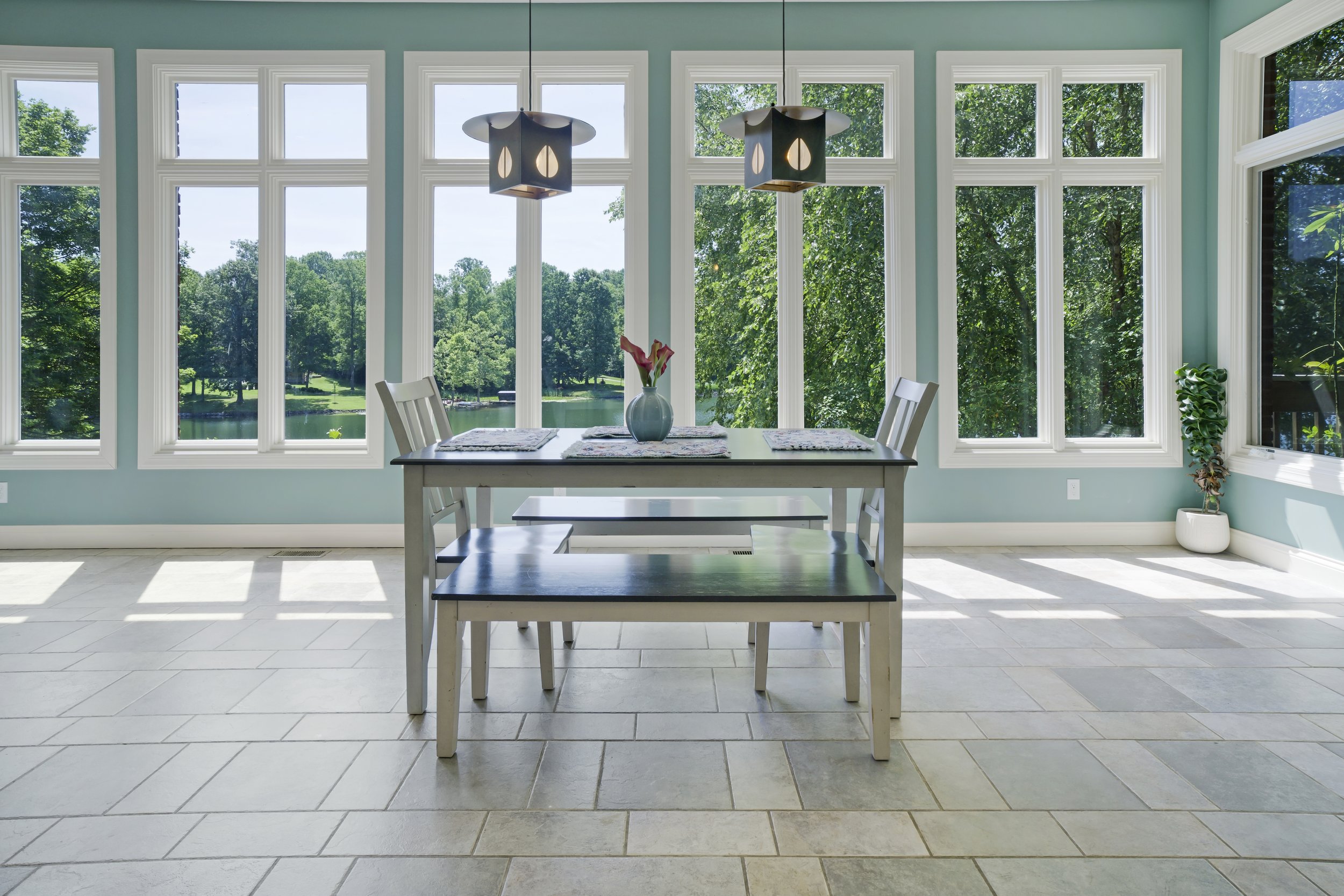 Bright dining room with large windows overlooking a green outdoor landscape, a table with a vase of flowers, and modern hanging light fixtures.