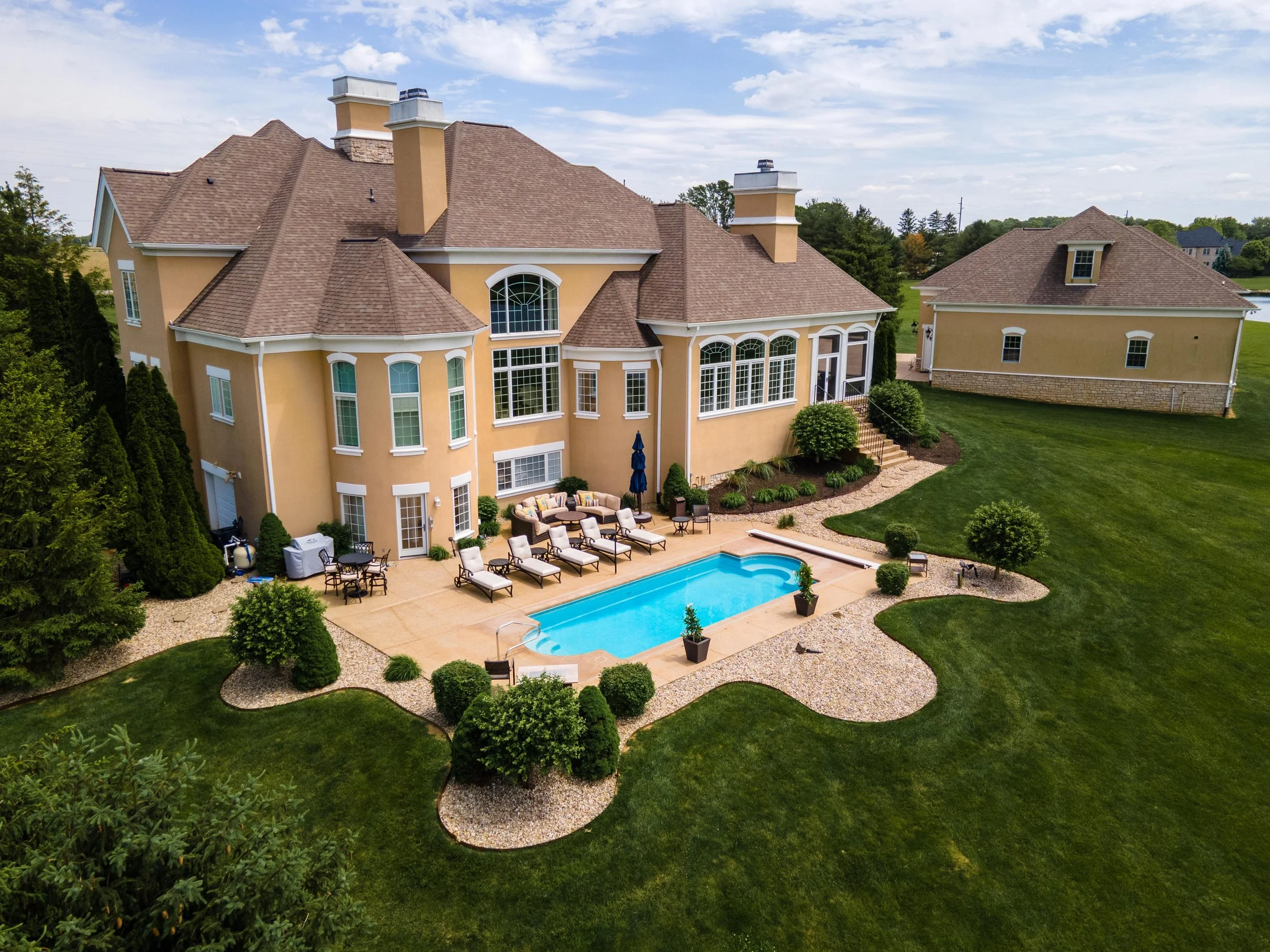 A large, yellow, two-story house with a backyard swimming pool, outdoor furniture, and lush green lawn, surrounded by trees under a partly cloudy sky.