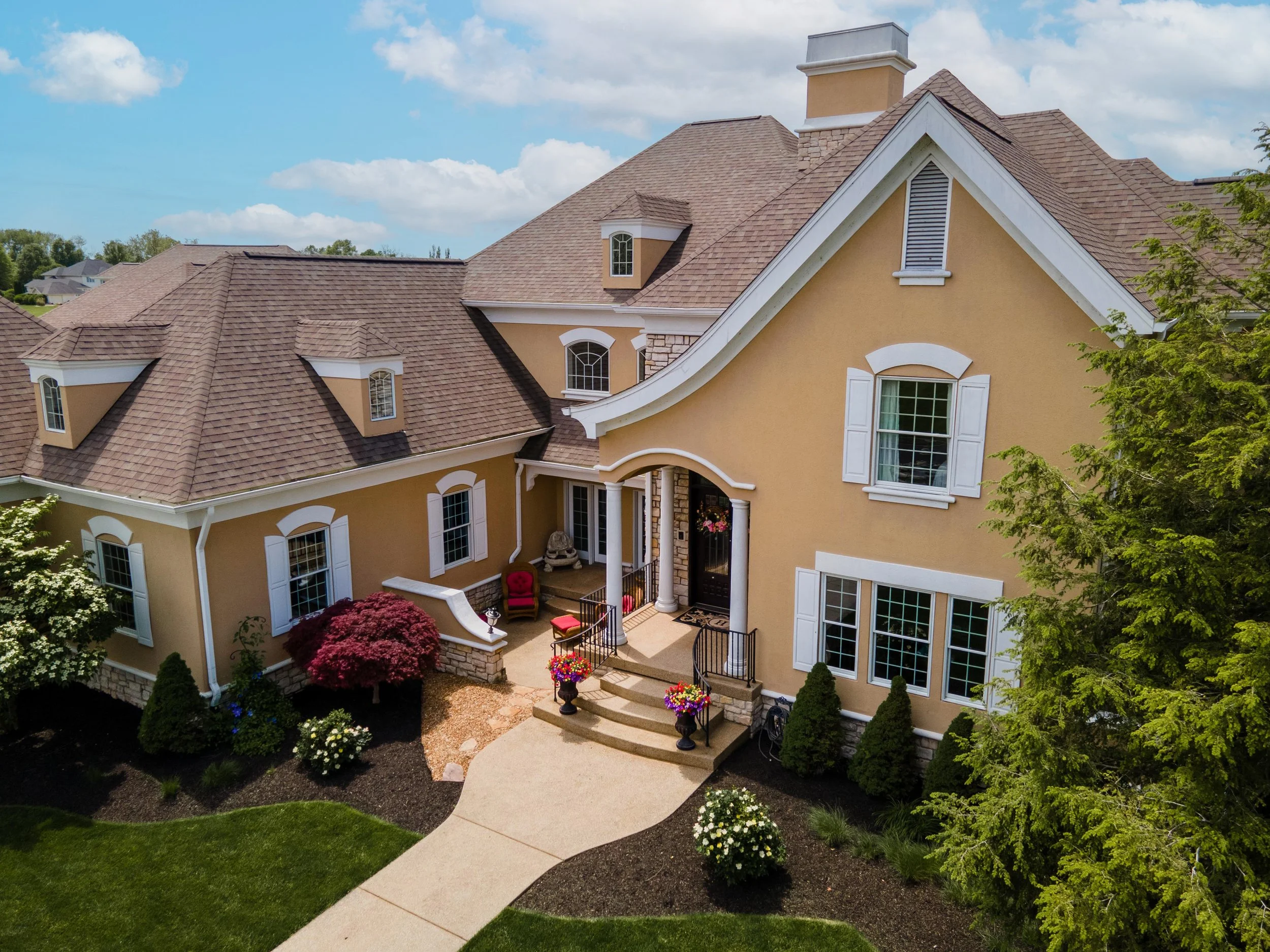 Front view of a large, yellow house with a brown shingle roof, white window shutters, and a landscaped yard with a curved concrete walkway, colorful flowers, and green shrubs.