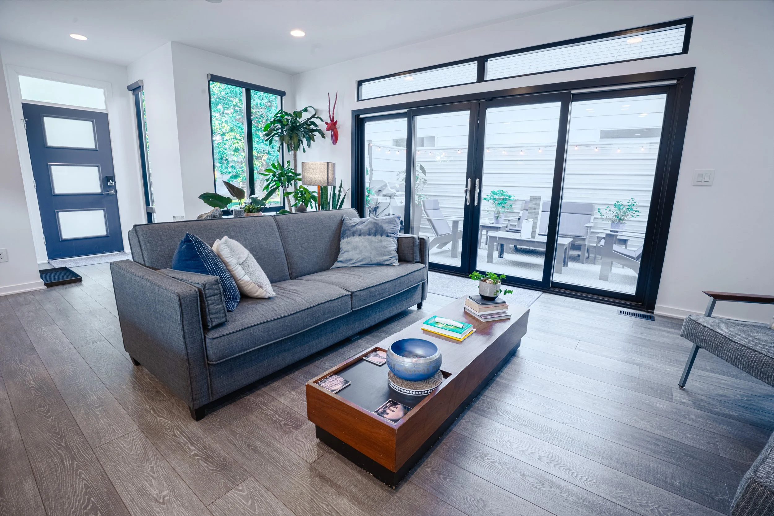 Living room with gray couch, plants near windows, glass sliding door leading to patio with table and chairs, wooden floor, and modern decor.