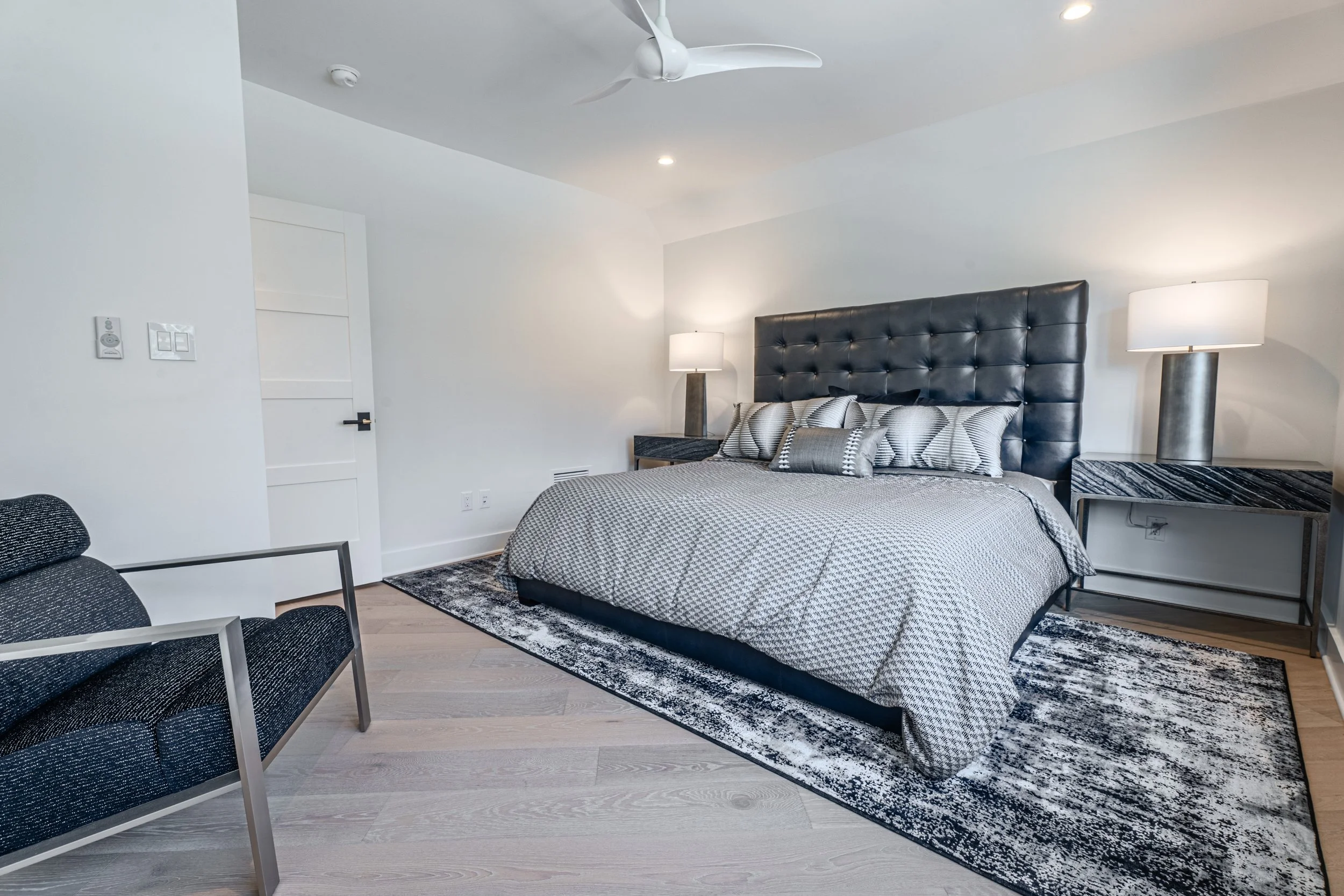 A bedroom with a large bed featuring a black tufted headboard, gray and white striped pillows, a gray and white patterned bedspread, two matching nightstands with lamps, a ceiling fan, and a rug on light wood floors.