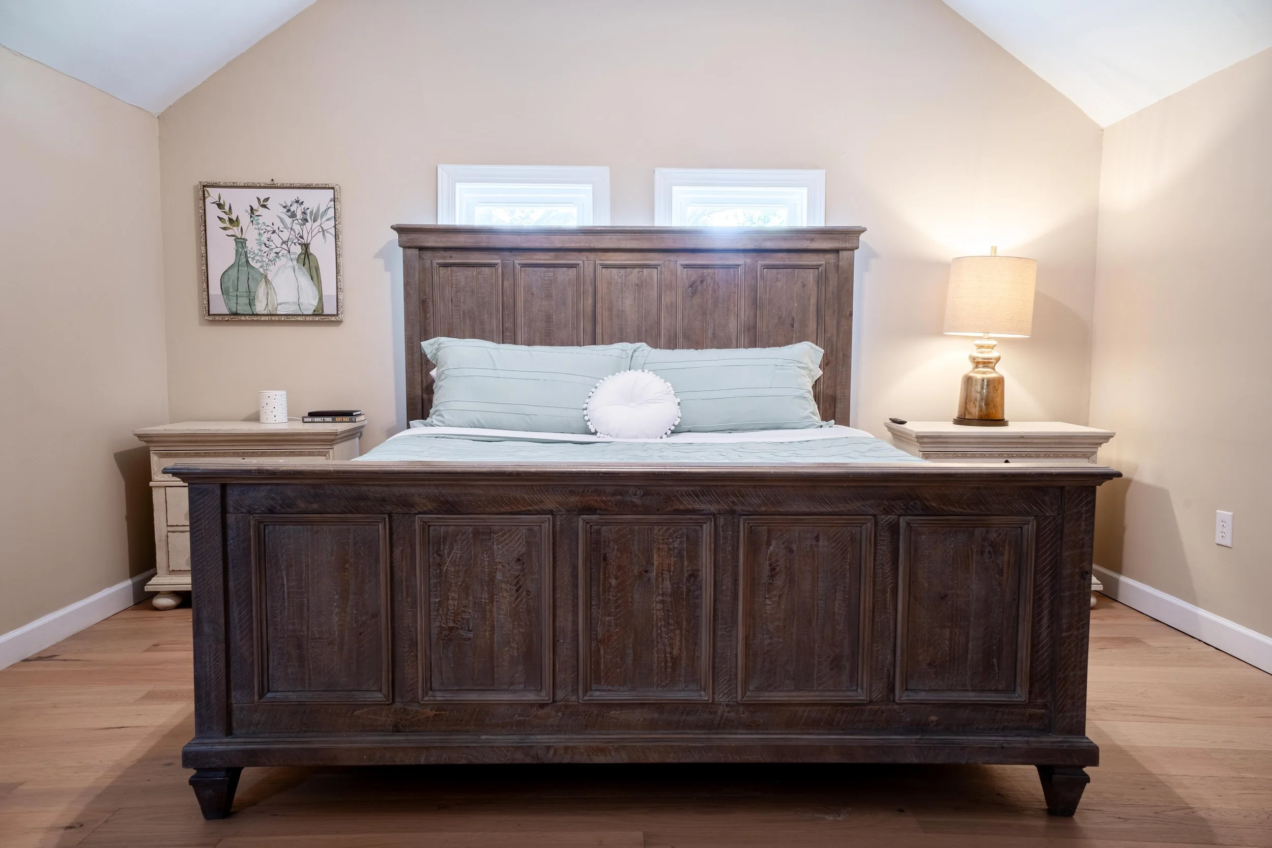 A bedroom with a wooden bed frame and a matching headboard, two small windows behind the headboard, a nightstand on each side, one with a lamp, a framed artwork of vases with floral illustrations on the wall, and a round decorative pillow on the bed.