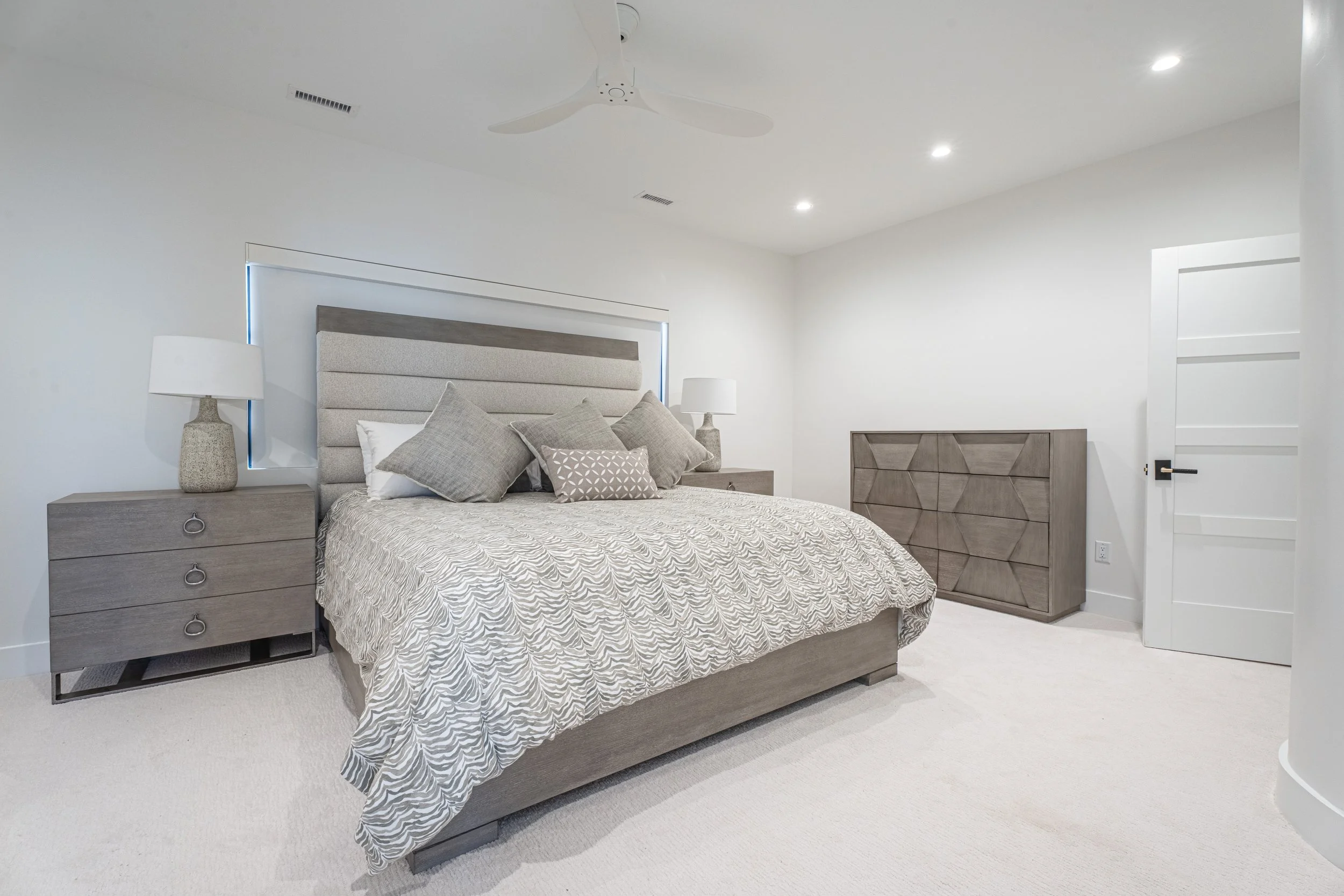 A bedroom with a bed featuring a padded headboard and a comforter with a wavy pattern. There are several pillows on the bed. On each side of the bed are matching nightstands with textured lamps. A dresser with a geometric pattern is against the wall, and a closed door is visible to the right. The room has recessed ceiling lights, a ceiling fan, and a window with a minimalist blind.