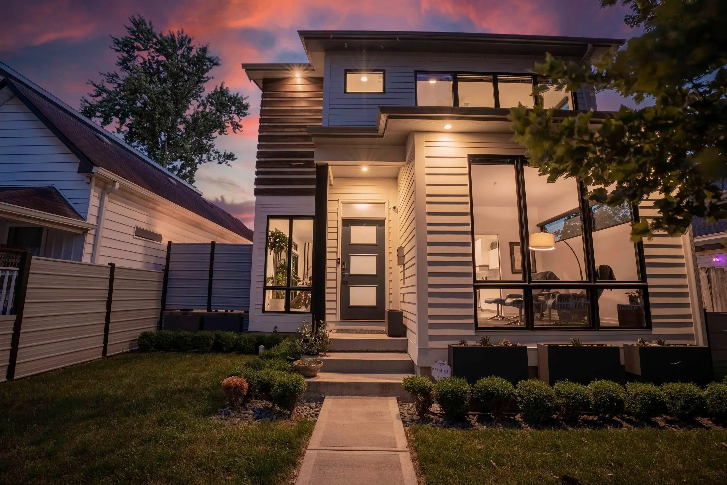 Contemporary two-story house with large glass windows and a small front yard with landscaped bushes, during sunset.