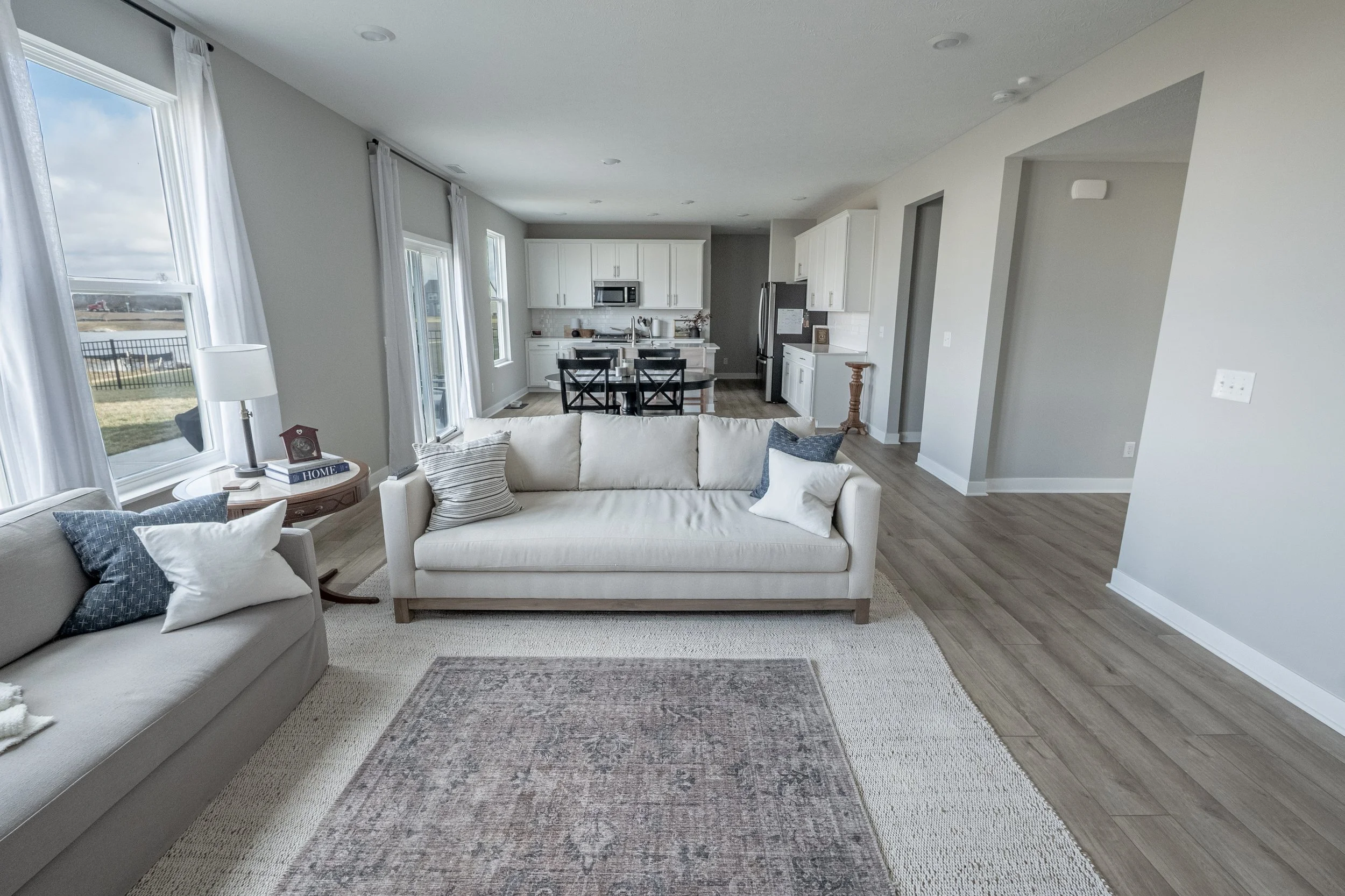 Open-concept living room with large windows, white sofas with decorative pillows, a wooden side table, and a view into the kitchen area with white cabinets, stainless steel appliances, and a dining table.