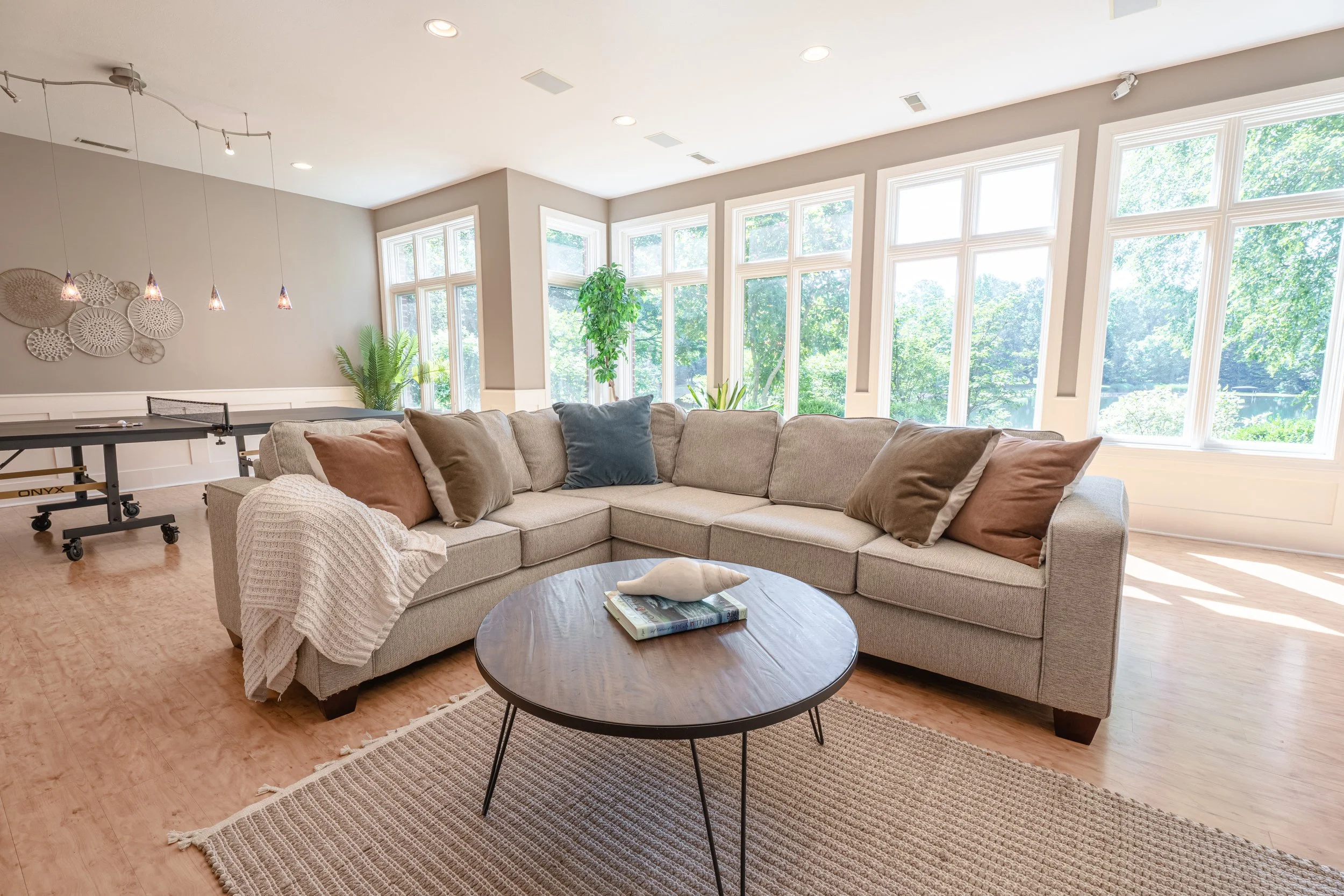 Living room with large sectional sofa, pillows, coffee table, plants, and large windows with green trees outside.