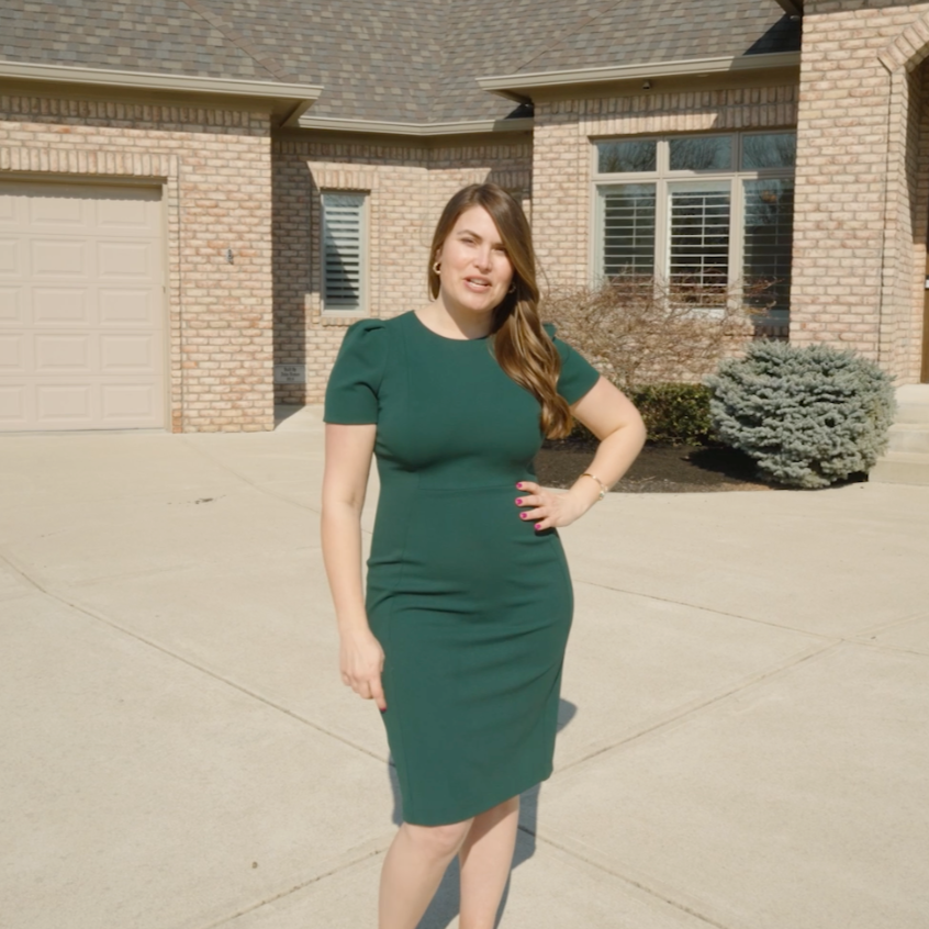 A woman standing outside in front of a brick house with a garage, wearing a green dress, with her hand on her hip and slightly squinting in the sunlight.