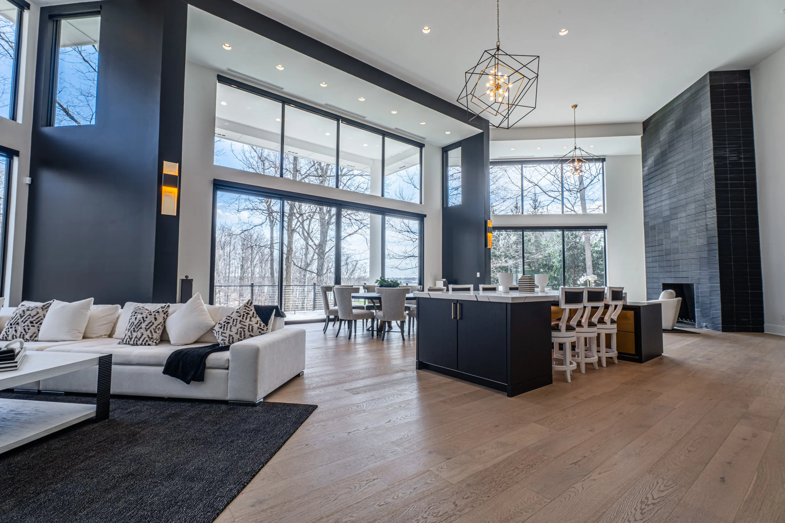 Interior of a modern, spacious living room with large floor-to-ceiling windows, a white sofa with patterned pillows, a wooden dining table with beige chairs, black and white kitchen island, contemporary geometric chandeliers, and wooden flooring.