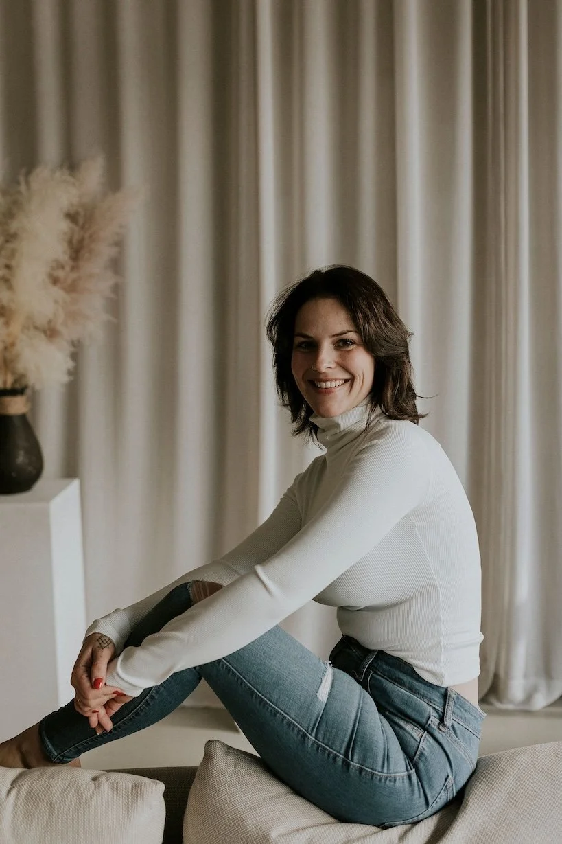 Une femme souriante avec des cheveux bruns, habillée d'un pull blanc et d'un jean, assise sur un canapé devant un rideau beige.