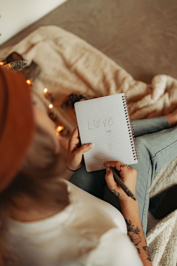 Une personne assise sur un canapé, tenant un carnet avec l'inscription 'Love' et un cœur, ainsi qu'un stylo, avec une couverture beige et des guirlandes lumineuses en arrière-plan.