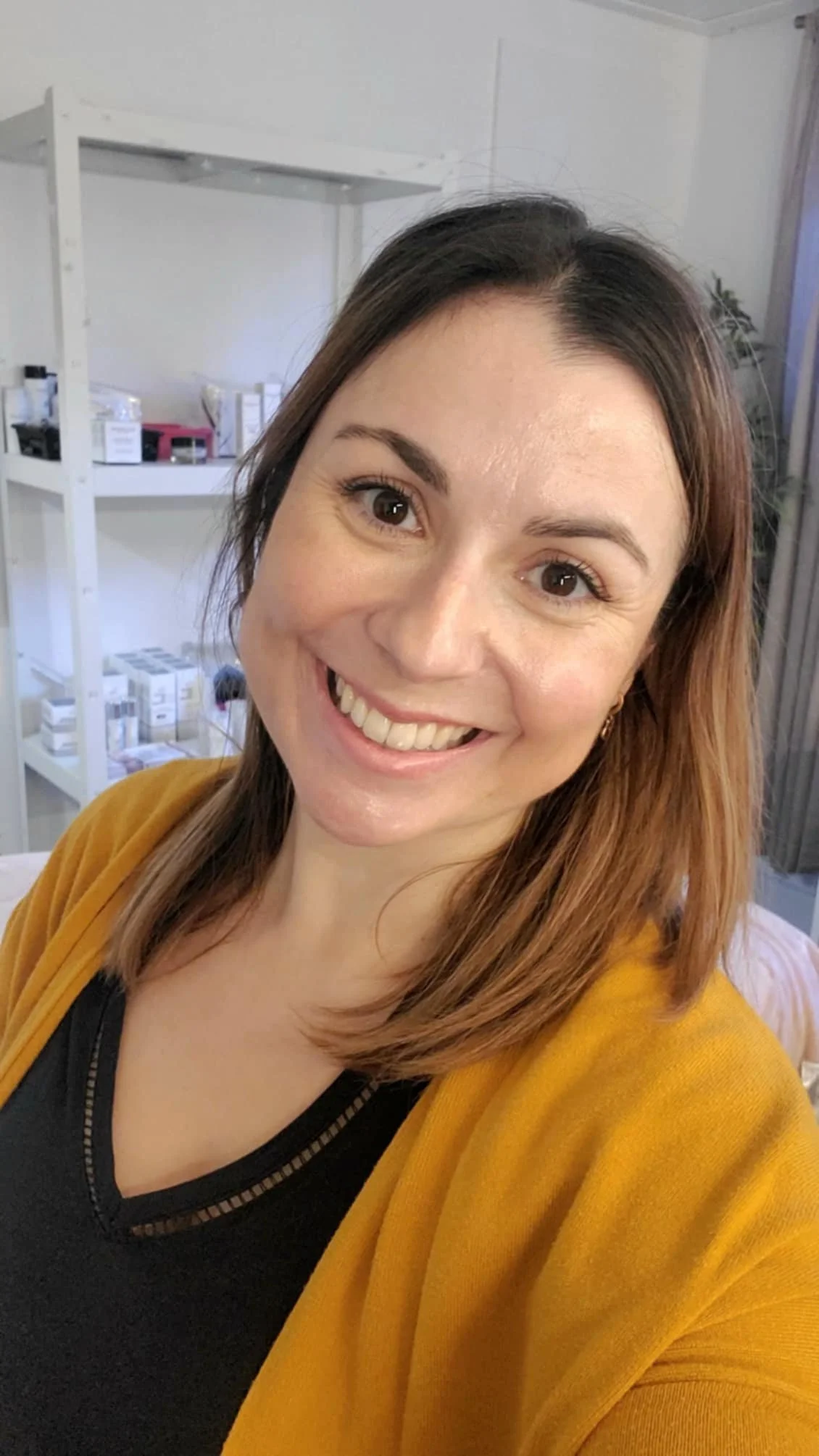 Une femme souriante avec des cheveux bruns et un pull jaune dans une pièce avec des étagères en arrière-plan.
