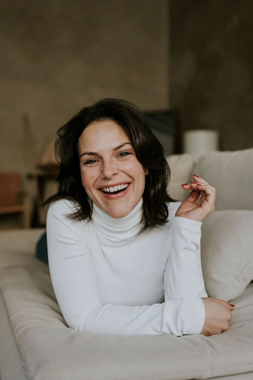 Une femme souriante avec des cheveux bruns foncés, portant un pull blanc, allongée sur un canapé beige dans un intérieur moderne. Elle a un nez percé et des ongles rouges.