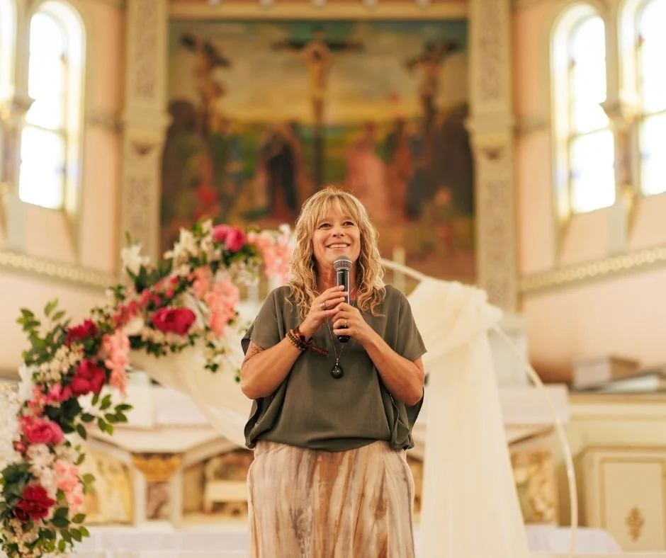 Femme tenant un microphone, parlant ou chantant devant une décoration florale dans une église ornée de vitraux et d'une grande peinture murale.