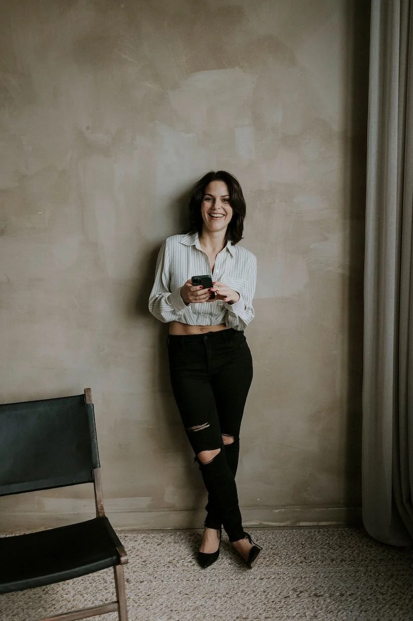 Une femme souriante qui regarde son téléphone, debout contre un mur beige, portant un chemisier blanc à rayures et un jean noir déchiré.