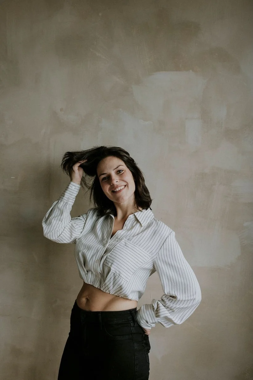 Une jeune femme souriante avec des cheveux bruns, portant une chemise blanche à rayures et un pantalon noir, se tenant devant un mur beige. Elle pose avec une main dans ses cheveux et l'autre sur la hanche.