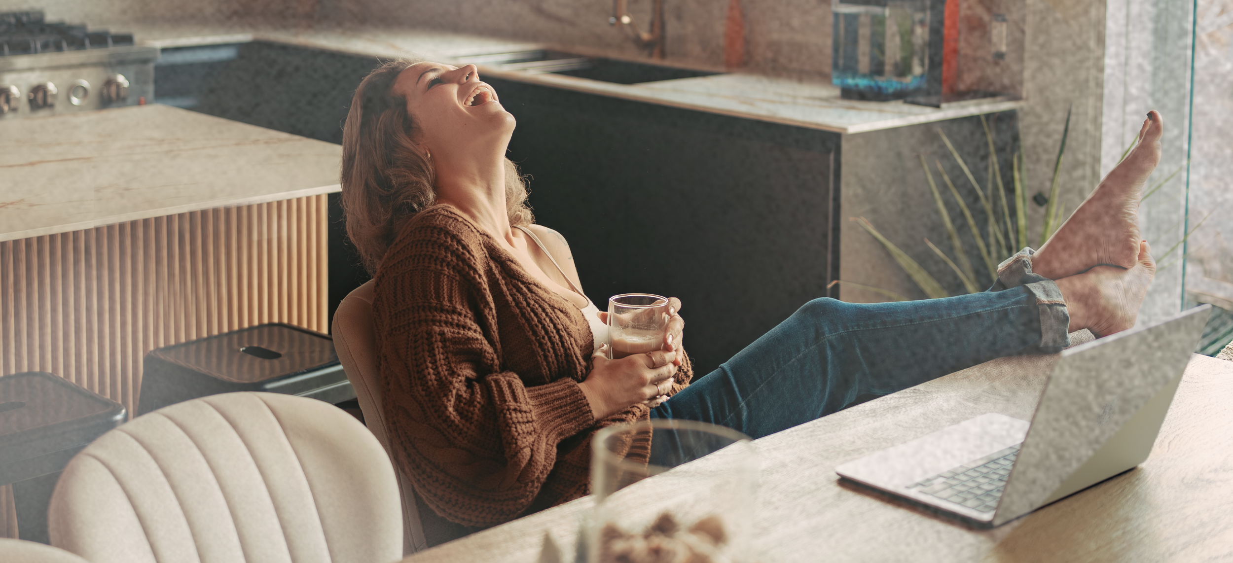 Femme souriante détendue assise à une table avec ordinateur portable, tenant un verre, jambes sur la table, dans une cuisine moderne