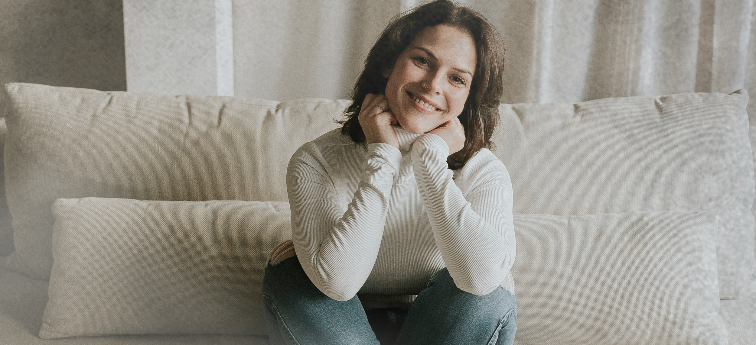 Jeune femme souriante avec un pull blanc et des jeans, assise sur un canapé beige, dans une pièce lumineuse avec des rideaux clairs en arrière-plan.