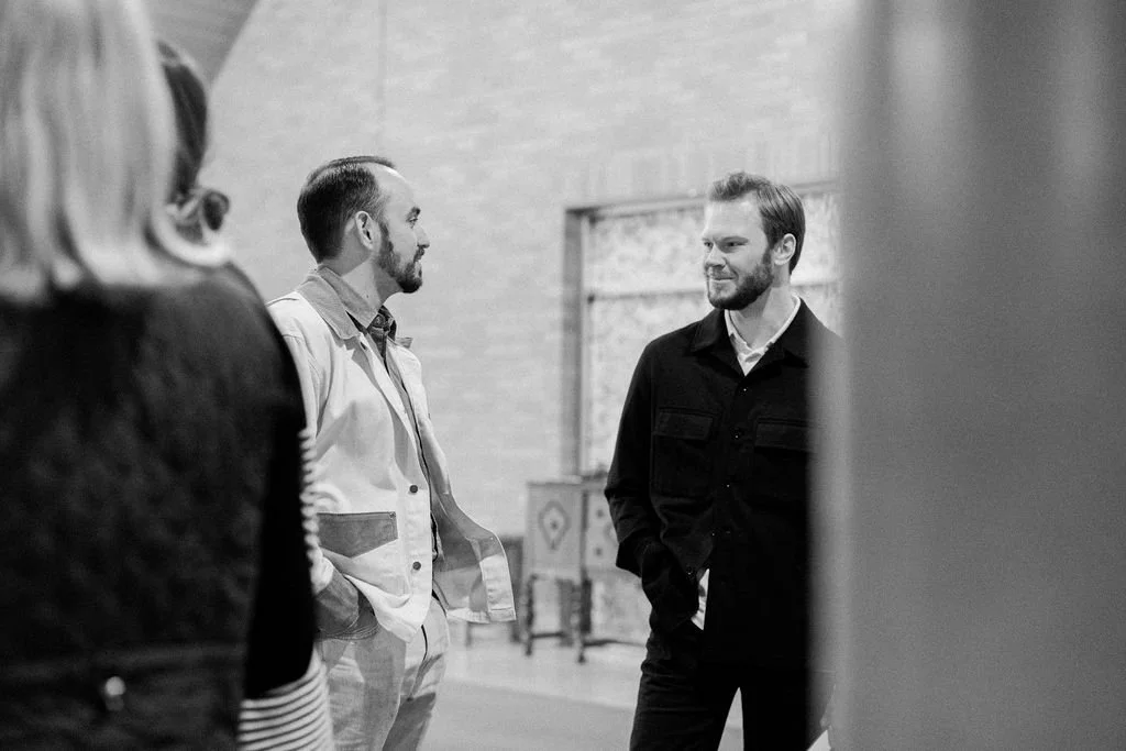 Two men are talking indoors, one wearing a light-colored jacket and the other in a dark jacket, with a woman partially visible in the foreground.