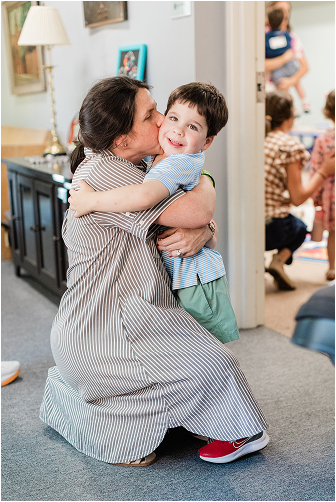 A woman in a striped dress kneeling and hugging a smiling young boy in a blue shirt in a living room, with other children playing in the background.