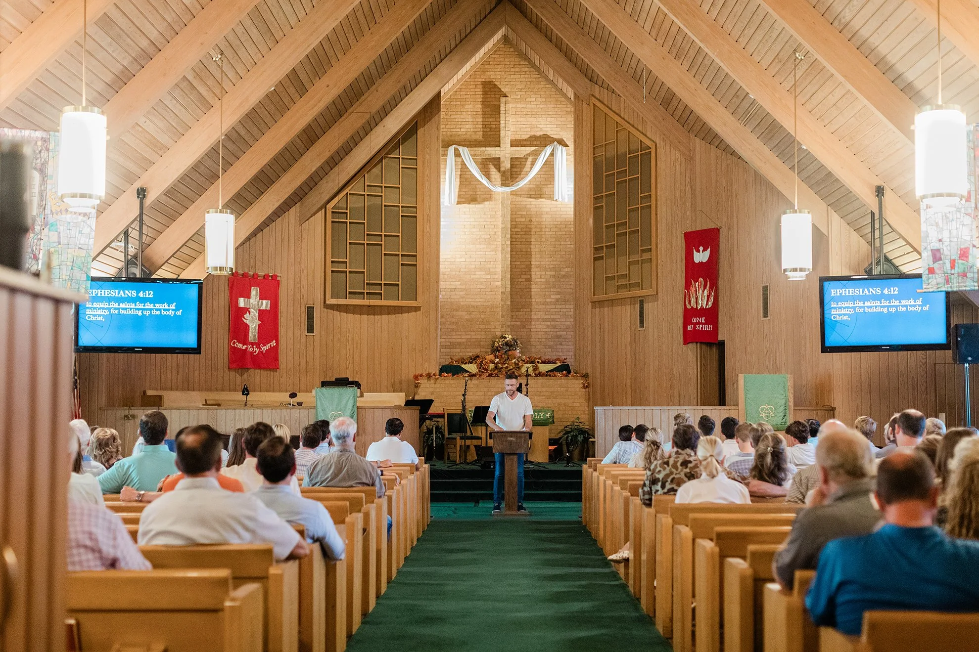 A congregation gathered in a church during a service, with a man speaking at the pulpit, a cross and decorations on the altar, banner with Christian symbols, and digital screens displaying Bible verses.