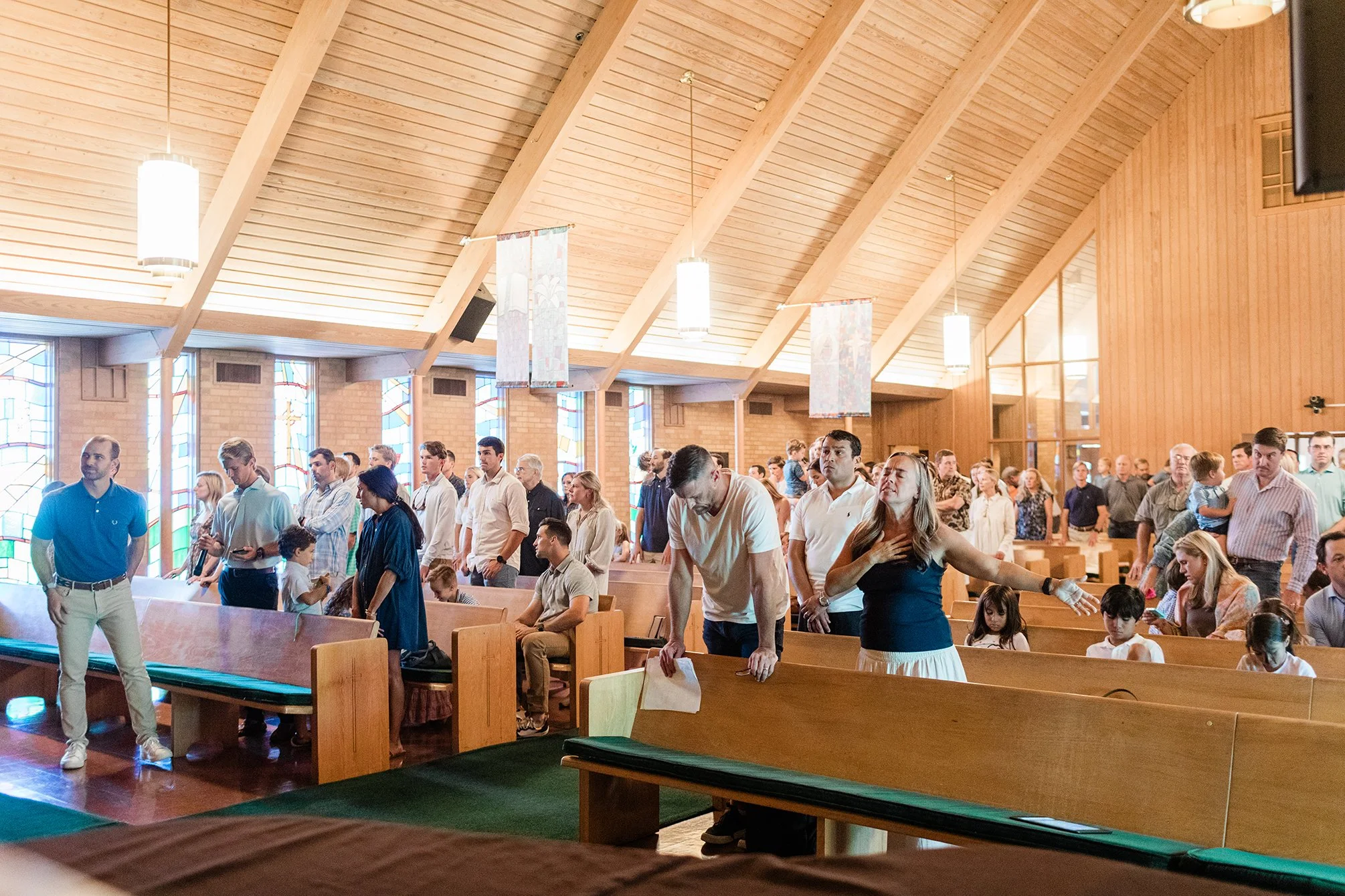 People attending a church service inside a wooden sanctuary with stained glass windows, some sitting, some standing, and a woman with closed eyes clasping her chest in prayer.