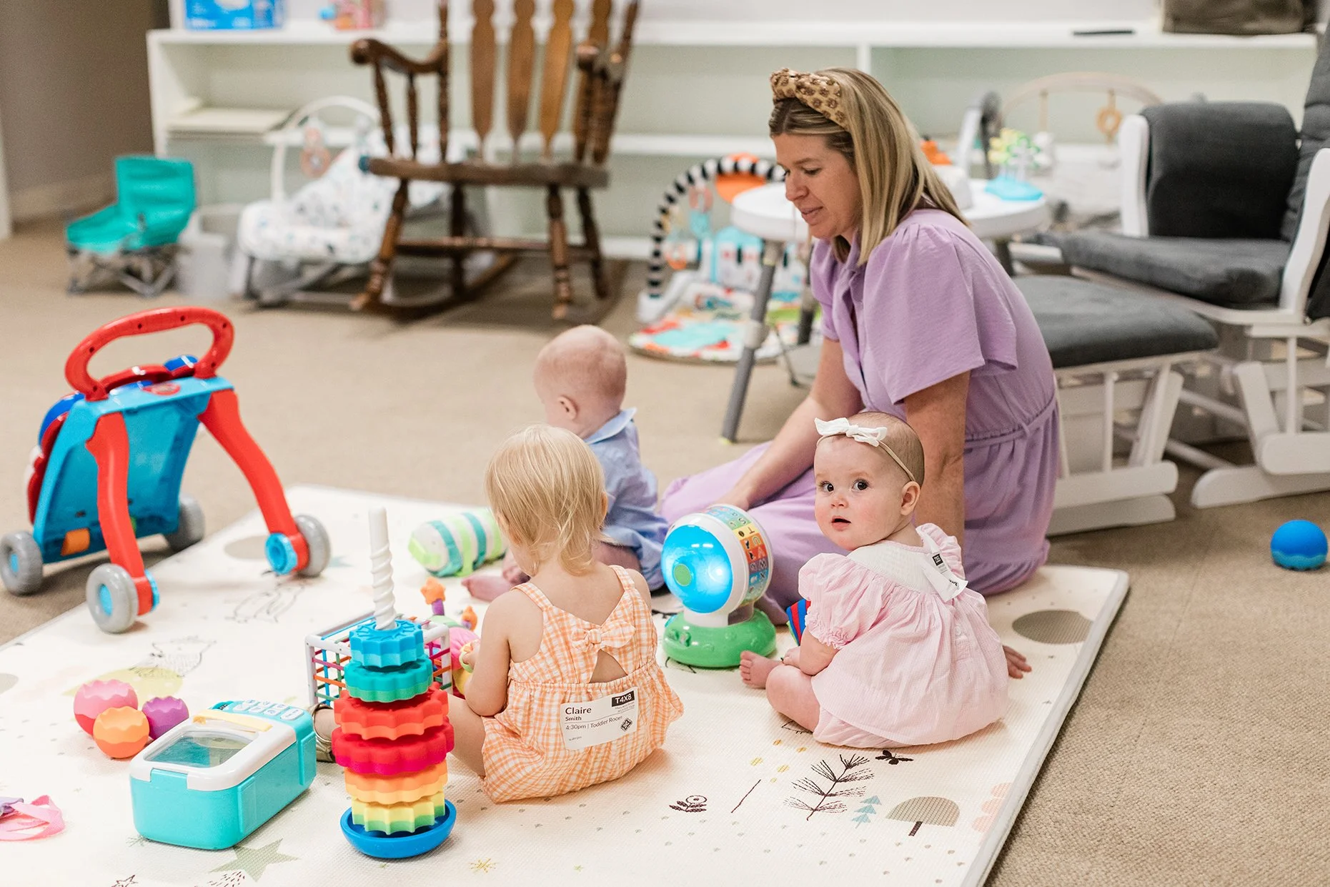 A woman with blonde hair wearing a lavender dress and a leopard headband sits on a play mat with three young children in a room with toys and furniture. The children, two girls and a boy, are surrounded by colorful toys, a red toy stroller, and a rob
