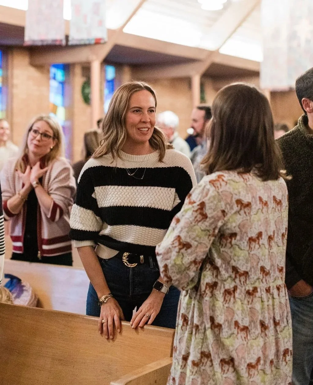 People gathered and talking inside a church with wooden pews and stained glass windows.