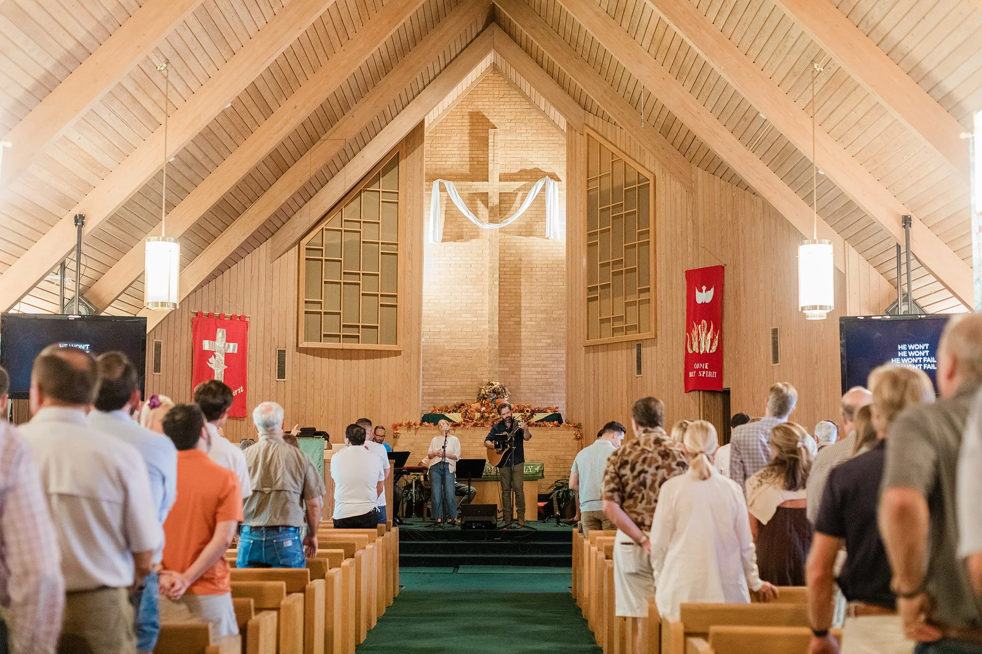 People gathered in a church with wooden interior, standing and facing the front where a band is performing on stage. There are banners with religious symbols on the walls, and a large cross decorates the back wall above an altar decorated with flower
