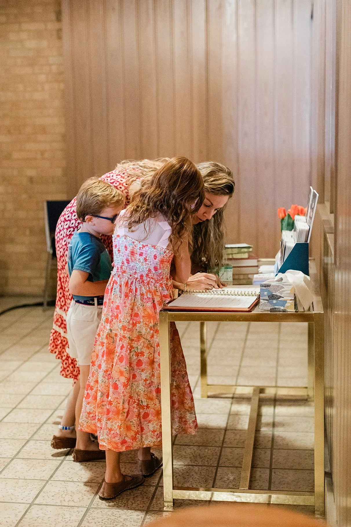 Three women and a young boy standing at a sign-in table, with the women writing in a guest book. The setting appears to be an indoor event, possibly a wedding or similar gathering.