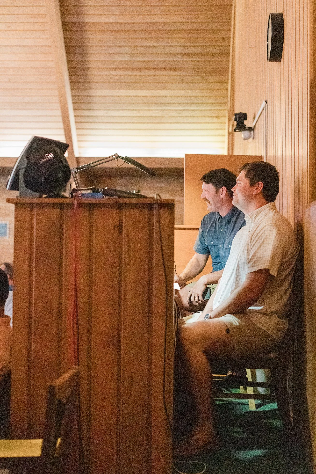 Two men sitting side by side in a room with wooden walls and ceiling, smiling and looking towards the left, possibly watching or listening to something.