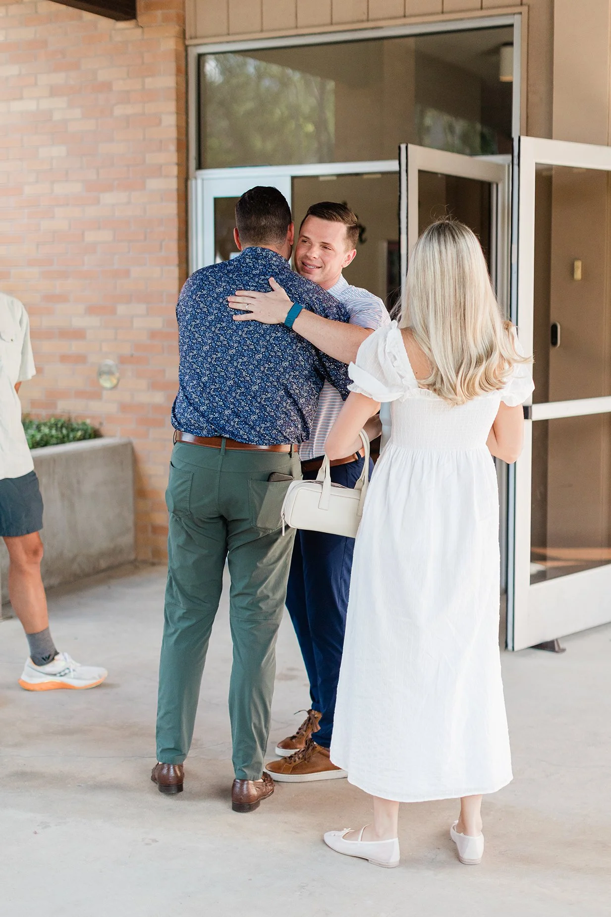 Two men and a woman greeting each other with hugs outside a building, with open glass doors behind them.