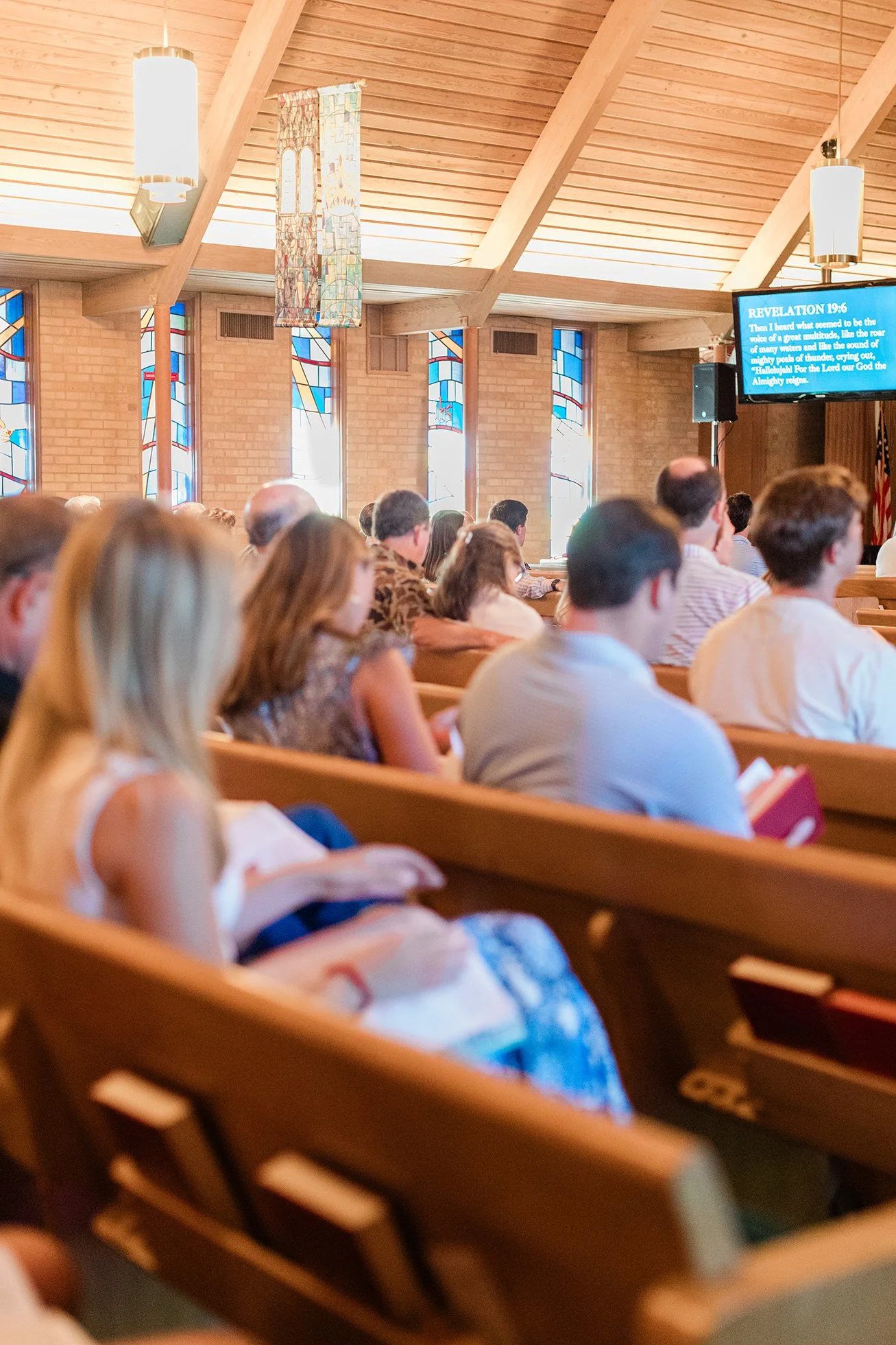 People sitting in pews inside a church, attending a service. There are stained glass windows, hanging light fixtures, and a screen displaying a Bible passage.