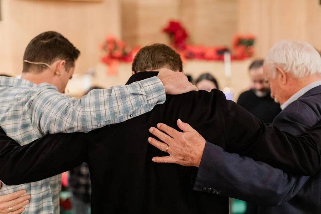 Three men with arms around each other's shoulders in a close circle during a prayer or moment of reflection, with a background decorated for Christmas.