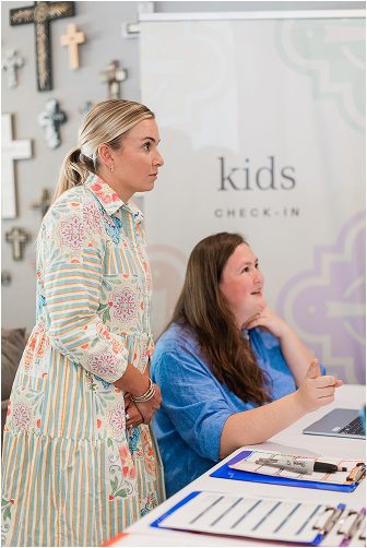 A woman standing and a girl sitting at a table with a laptop and notebooks, with crosses on the wall behind them, at a kids check-in area.