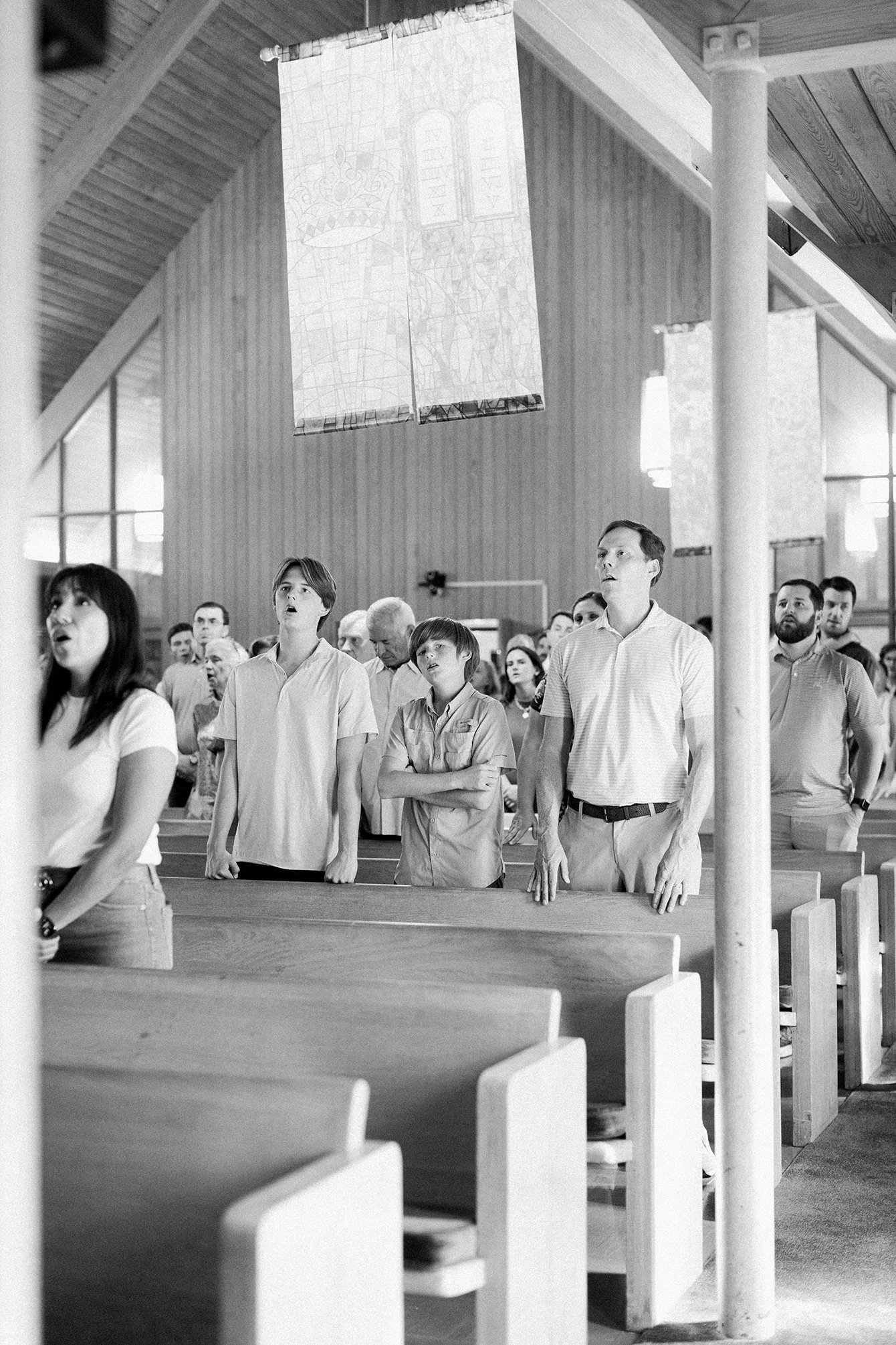 A congregation singing during a church service in a modern wooden church interior, with people standing and some having their eyes closed.