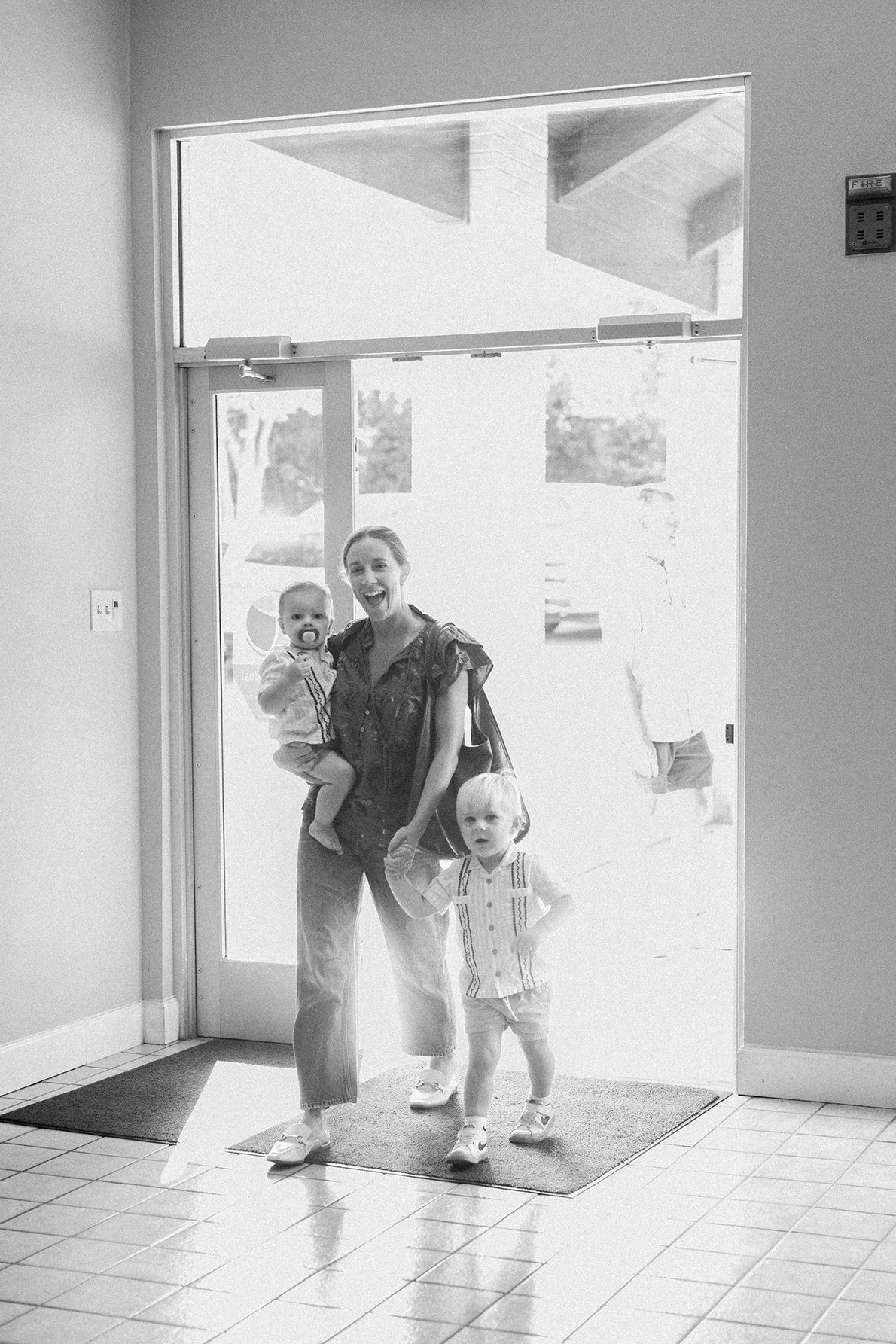 A woman and two young children entering a building through a glass door, with the woman smiling and holding a child, while the other child walks in front.