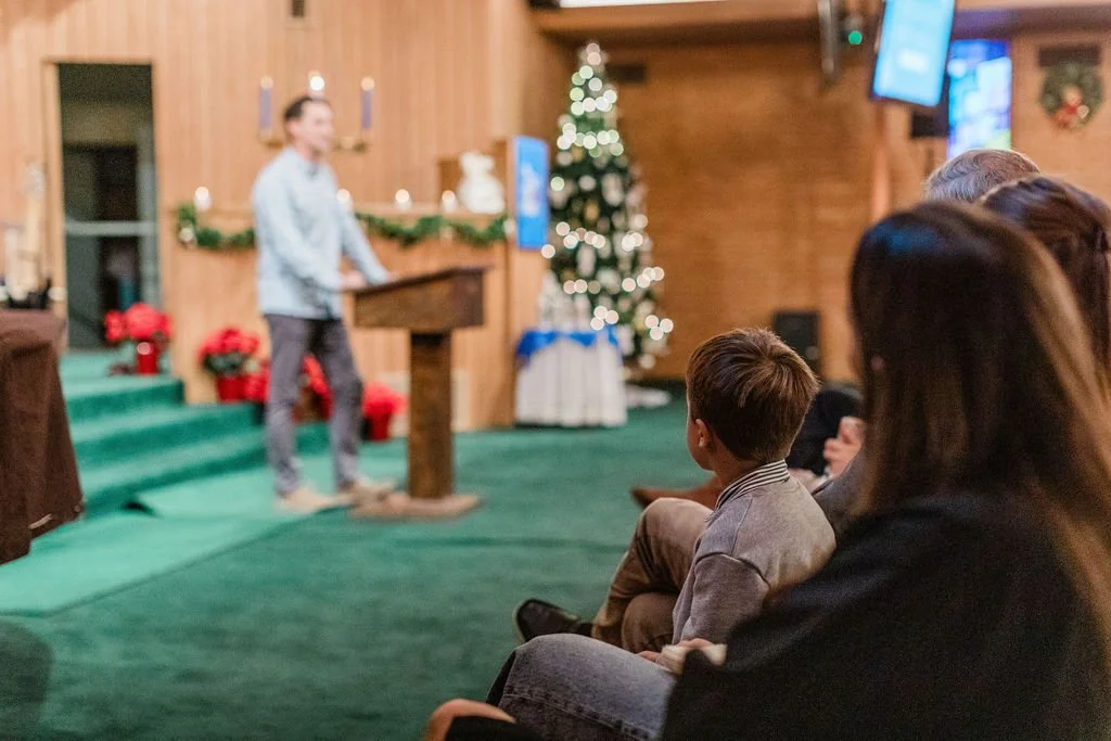 People sitting in a room watching a person speaking at a podium, decorated with Christmas trees and holiday decorations.