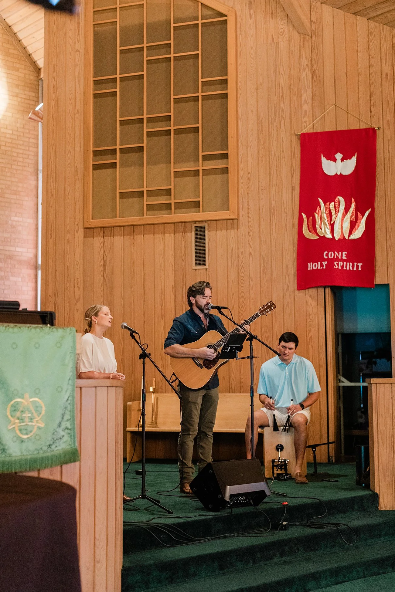 A man playing an acoustic guitar and singing into a microphone on stage, with a woman nearby and a young man sitting on a cajón behind him, inside a wooden-paneled church.