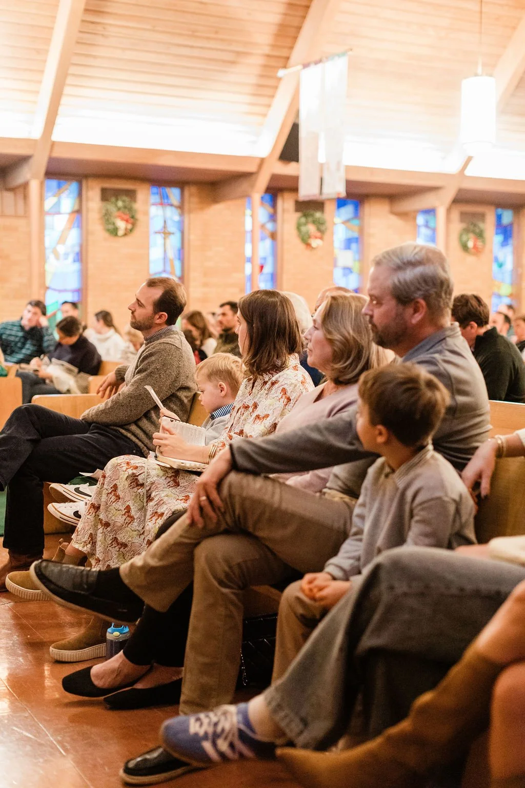 People sitting on pews inside a church, attending a service or event, with stained glass windows and wreath decorations in the background.