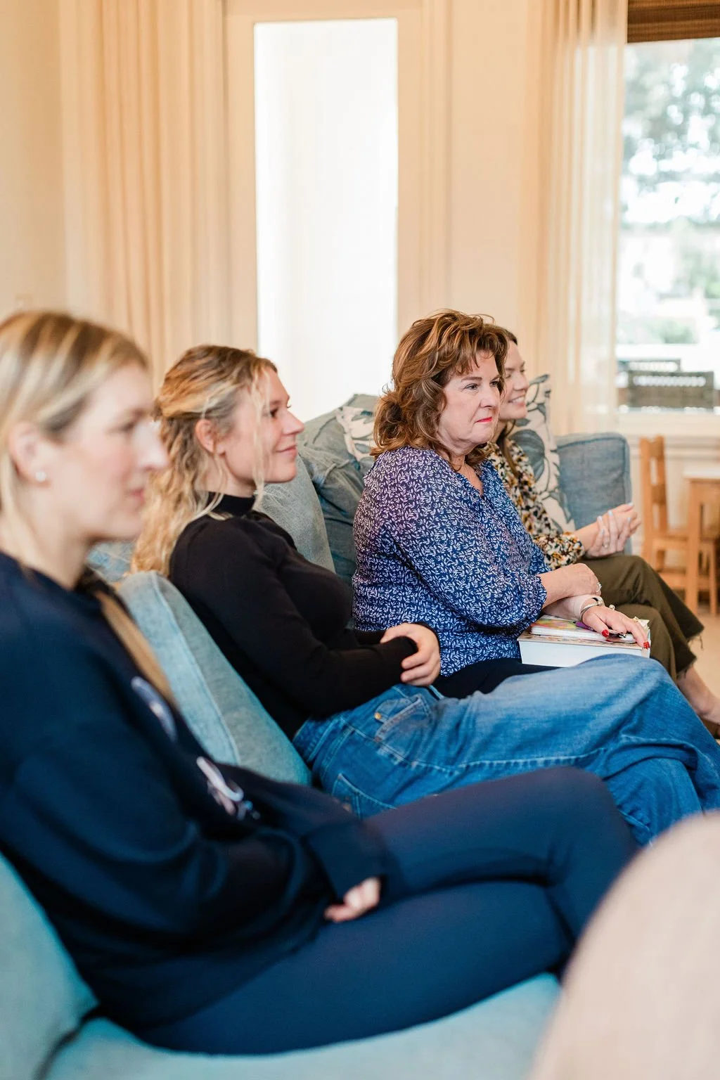 Four women sitting on a couch attentively listening at a gathering in a bright living room with large windows.