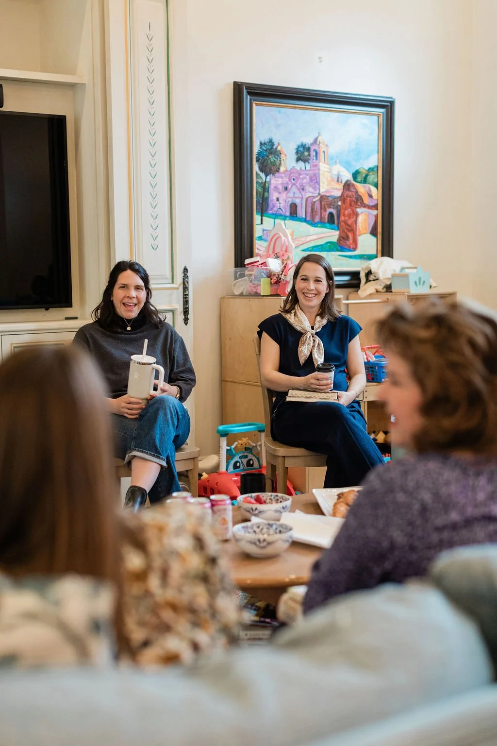 Group of women sitting and smiling around a table with food and drinks, with two women seated on chairs against the wall in the background, one holding a mug and the other a coffee cup.