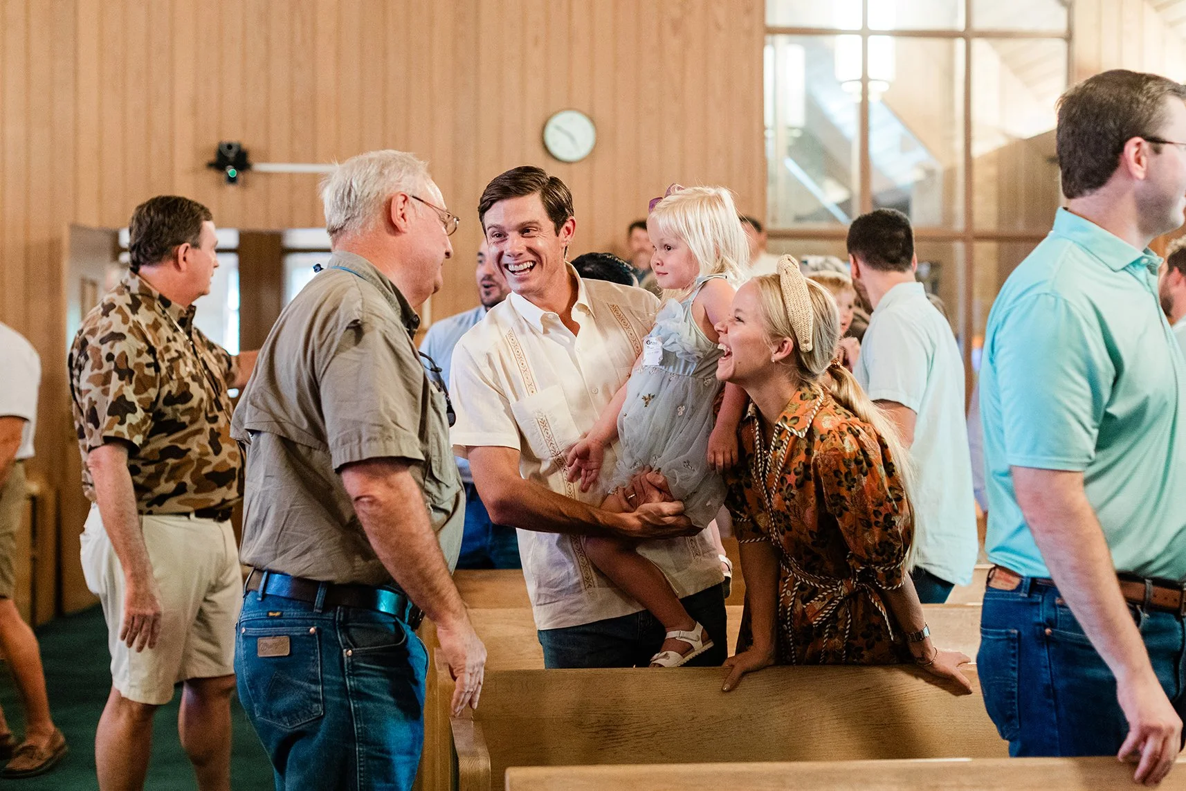 A group of people in a church, smiling and talking. Two men are in the foreground, one holding a young girl dressed in a light blue dress. There is a woman leaning on a pew, laughing, with others in the background.